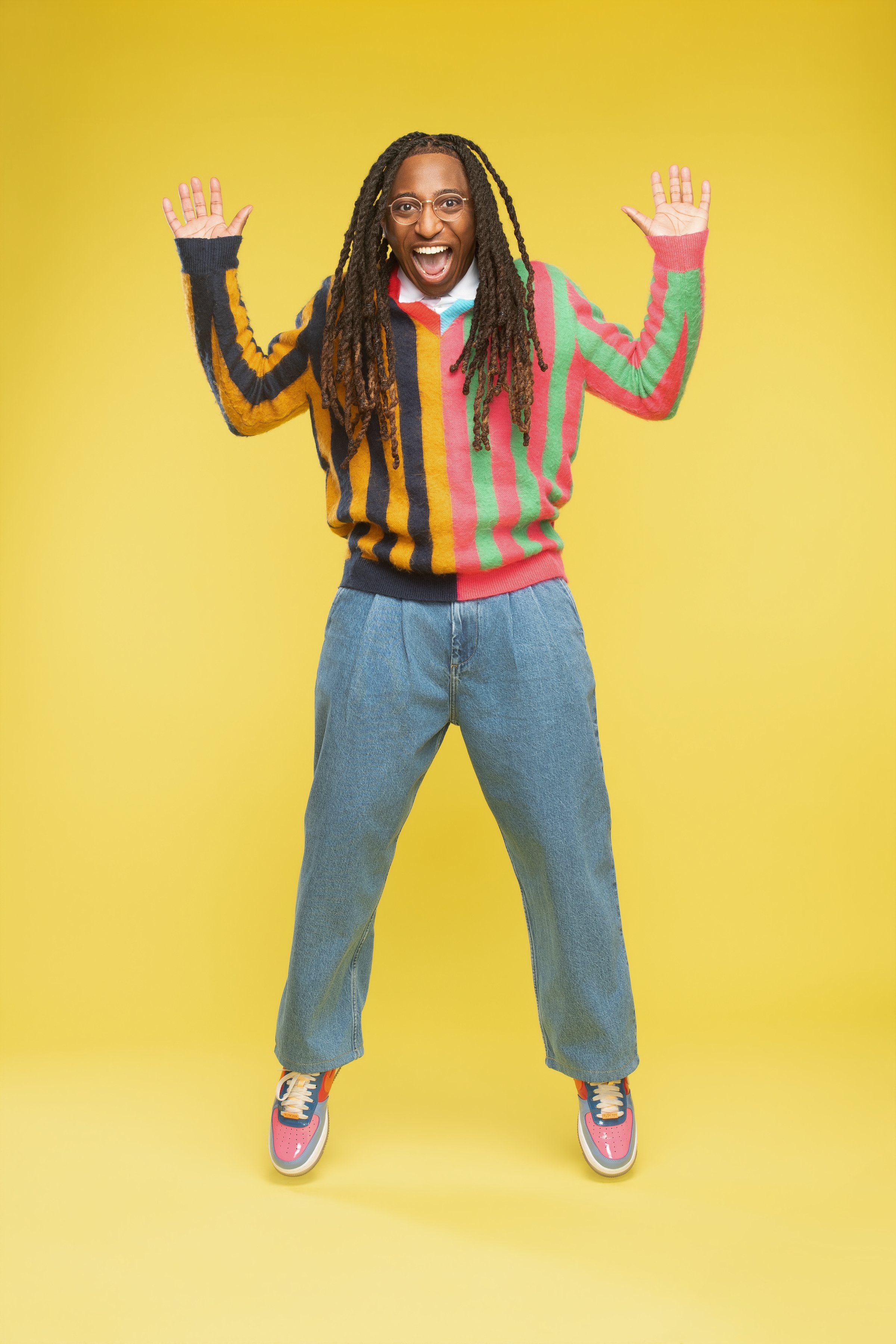 A person with dreadlocks wearing a colorful striped sweater, blue jeans, and sneakers, smiling with arms raised against a yellow background.