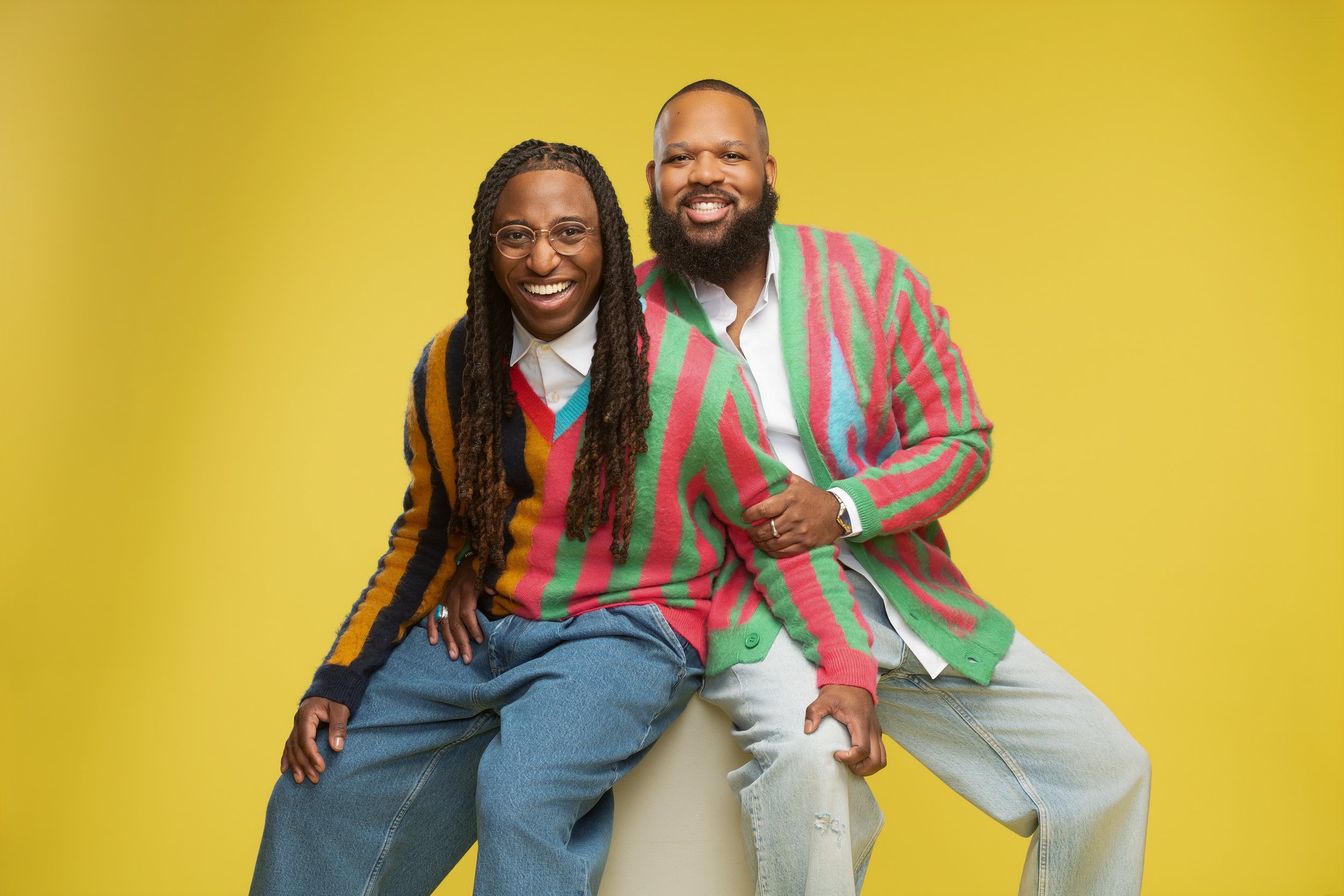 Two smiling people, one with dreadlocks and glasses and the other with a beard, wearing colorful sweaters, sitting against a yellow background.