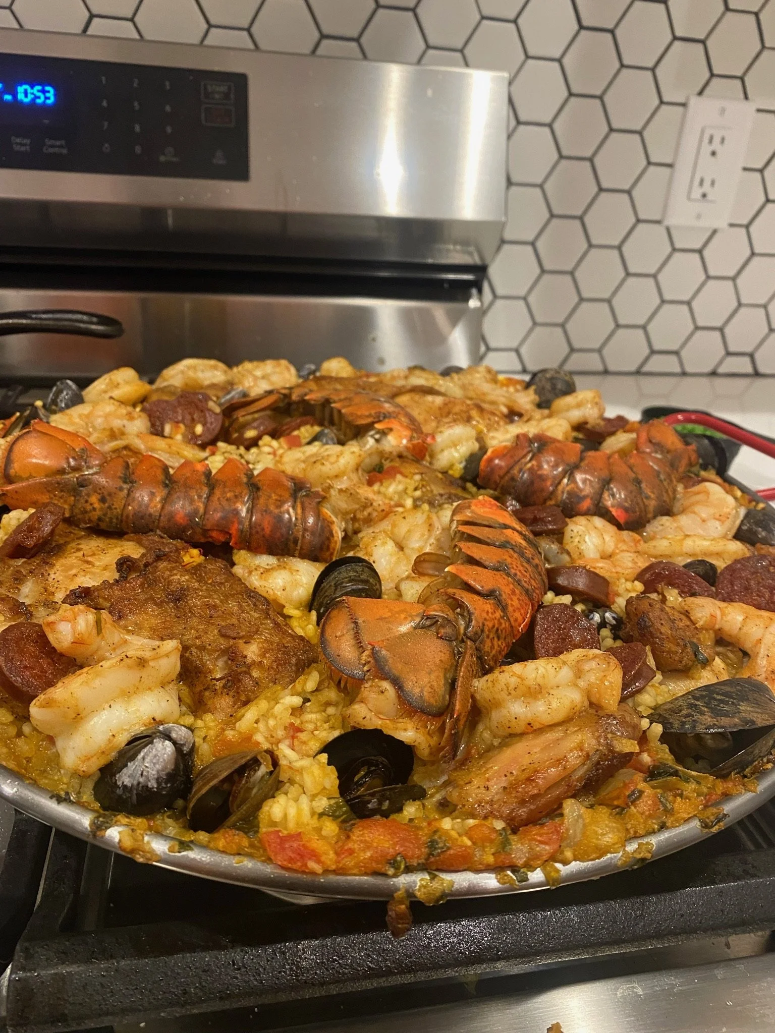 Seafood paella with lobster, shrimp, mussels, clams, and chorizo in a large skillet on a stove, against a white hexagon tile backsplash.