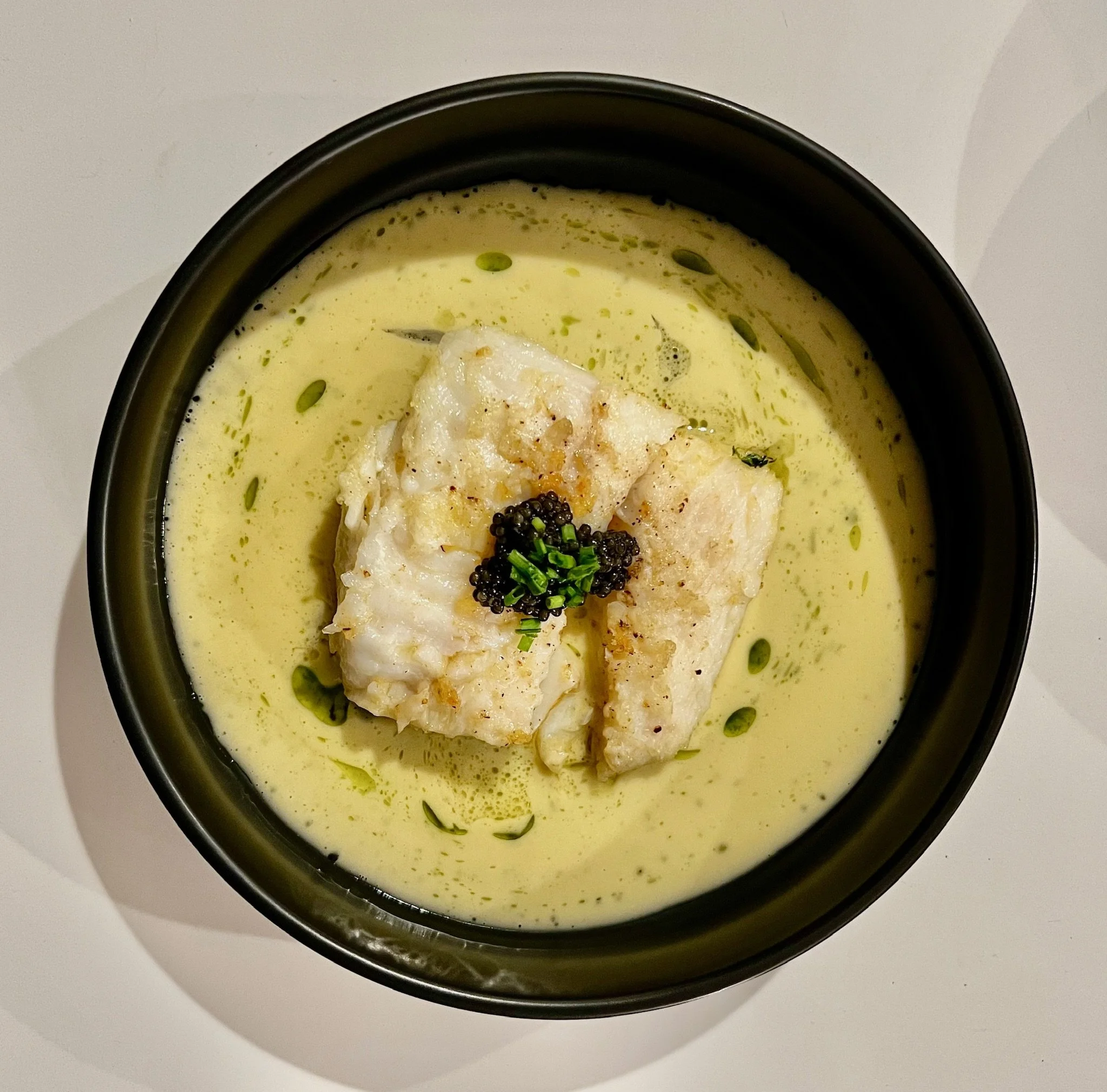 Bowl containing a creamy soup garnished with caviar, chopped chives, and a dollop of white fish fillet, with a white countertop background.