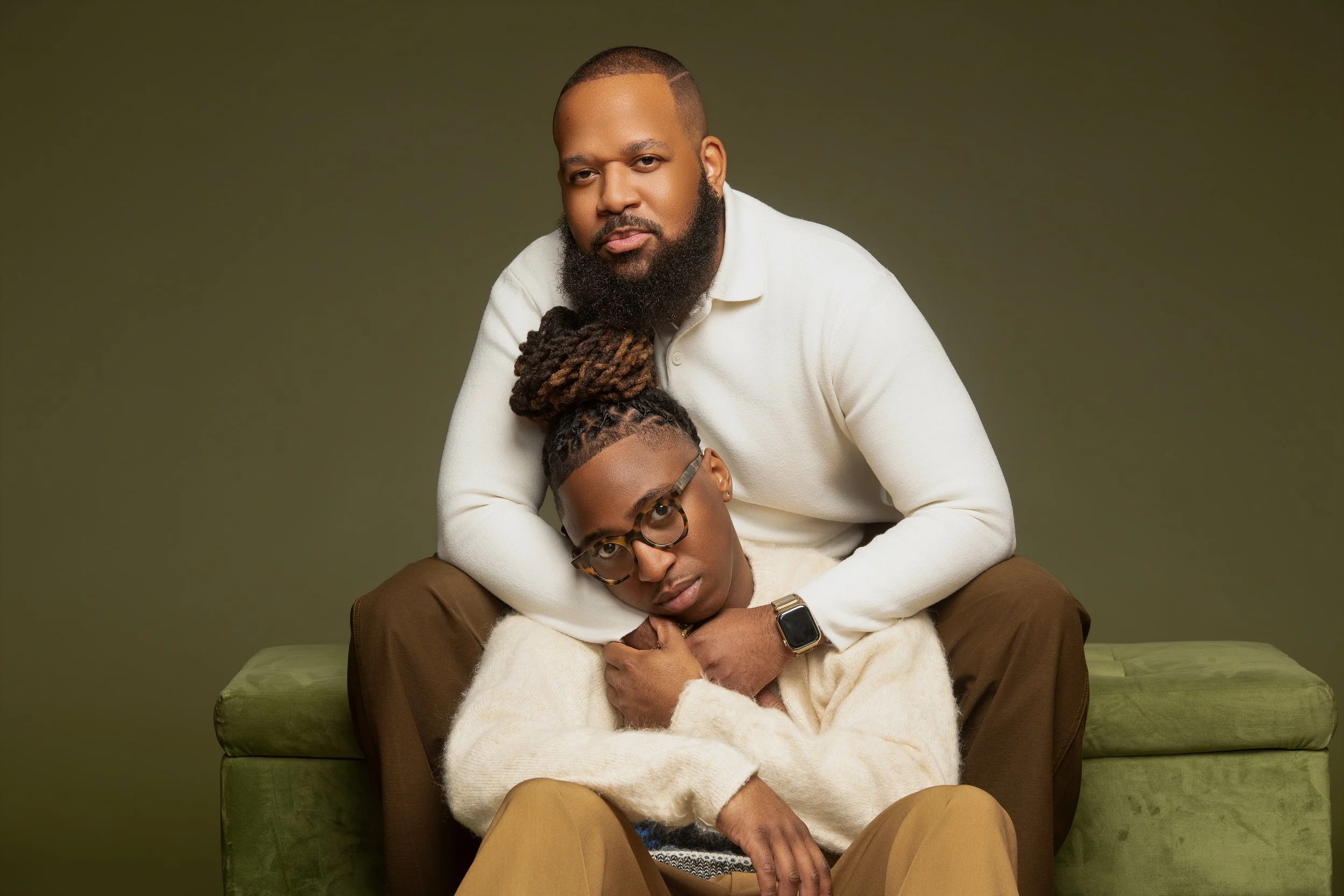 A man with a beard and a woman with glasses sitting on a green couch, with the man embracing the woman from behind, against a plain olive-green background.