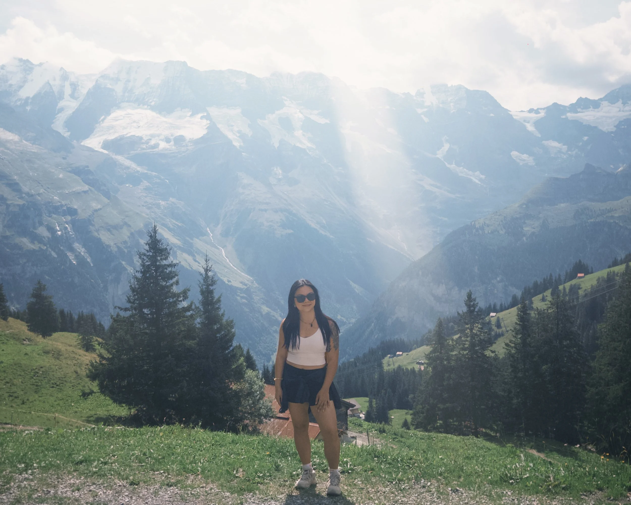 Melissa with long dark hair and sunglasses standing in a green, hilly landscape with mountains in the background, partially covered by clouds, and a small house on the right side | Human Bean | Therapy Space for you to be Human