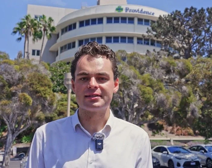 A young man with dark curly hair and wearing a white shirt stands outdoors in front of a hospital building with a sign that reads "Providence" and some parked cars. The background includes trees and clear blue sky.