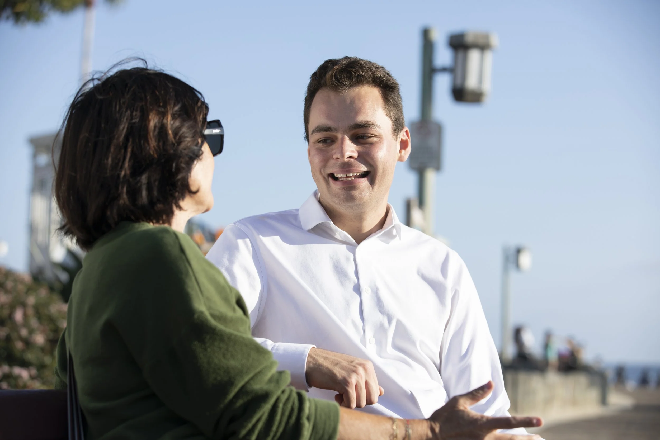 A young man and an older woman are sitting outdoors, engaged in conversation, with a clear blue sky and communication towers in the background.