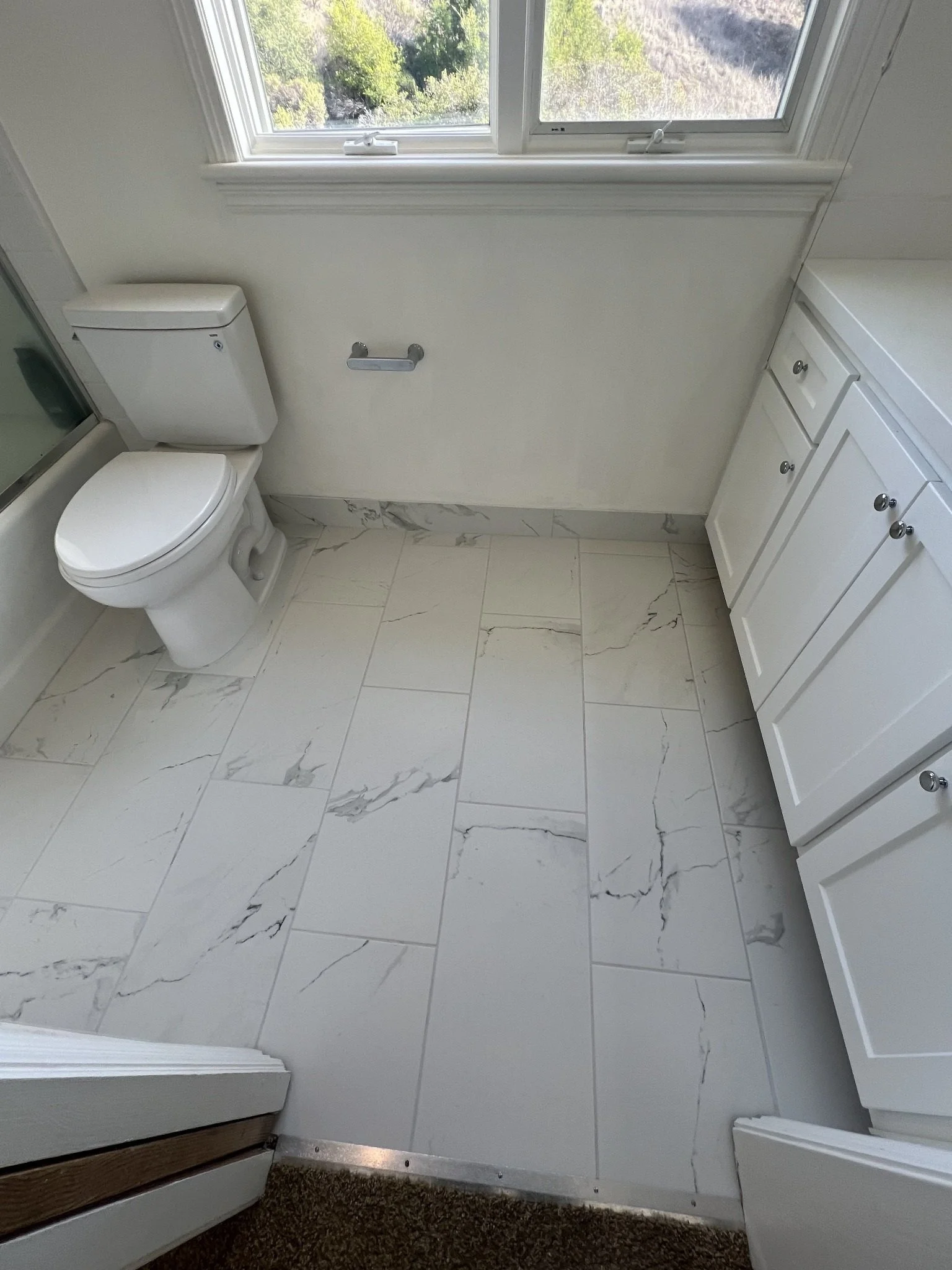 A bathroom with a toilet, white cabinetry, a window above the toilet, and marble tile flooring.