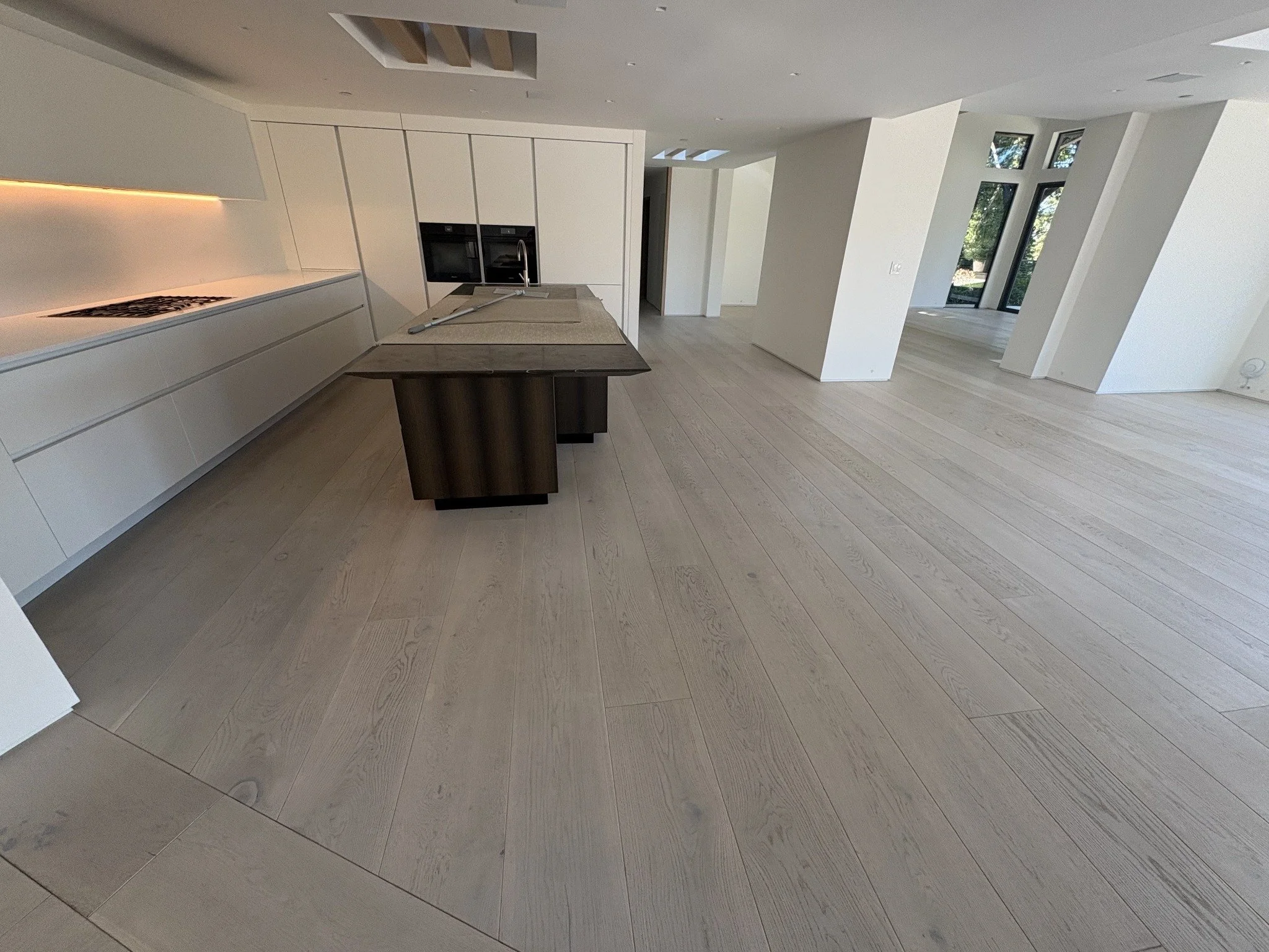 Modern open-concept kitchen and living space with white walls, light wooden floors, large windows, and a minimalist island with a sink.