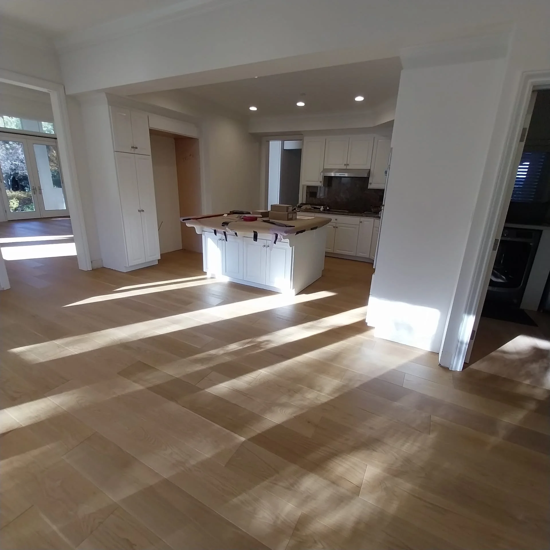Interior view of a modern kitchen with white cabinets, a central island, and hardwood flooring. Sunlight streams through a large window, casting shadows on the floor.