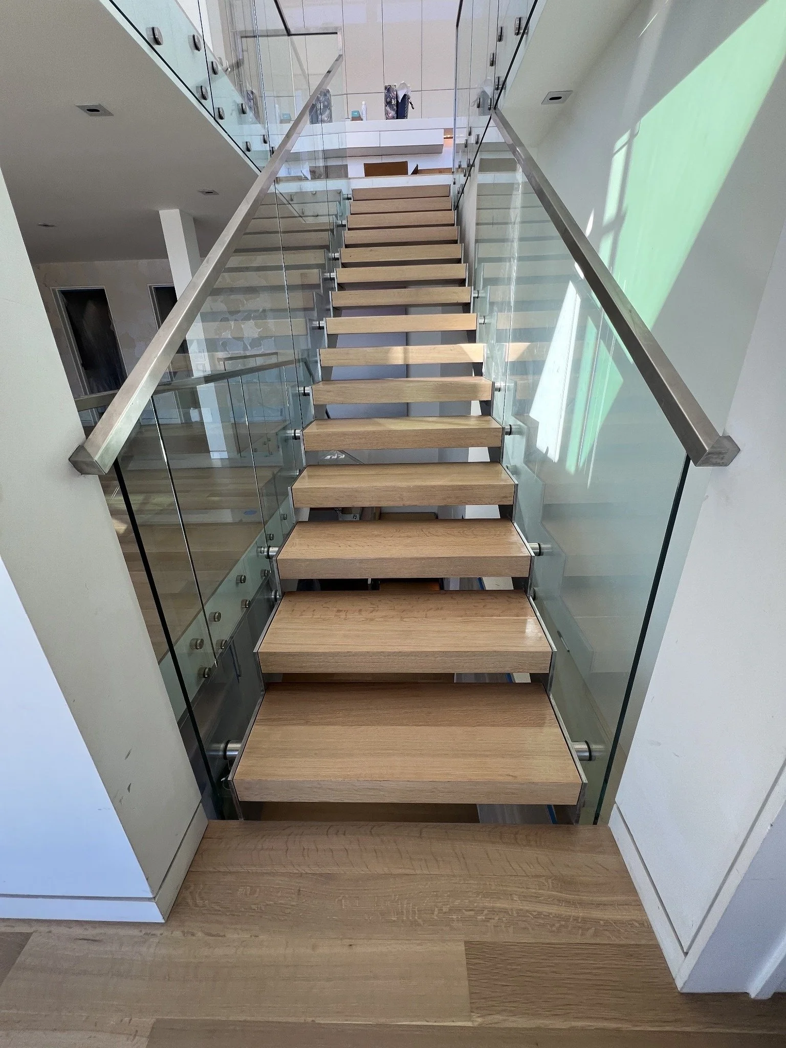 Interior view of a modern staircase with wooden steps, glass railings, and white walls, leading to an upper level with a reception desk and decorative items.