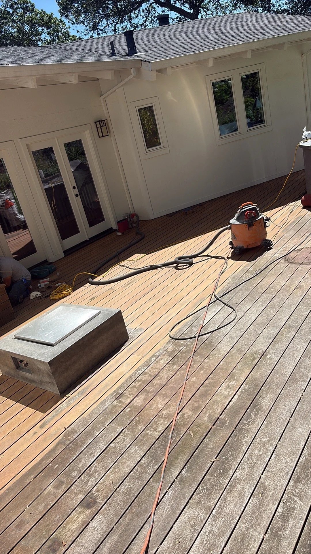 View of a backyard wooden deck with construction tools and equipment, including a wet vacuum, extension cords, a fire extinguisher, and a vent or chimney on the deck, with a house wall featuring windows and doors.