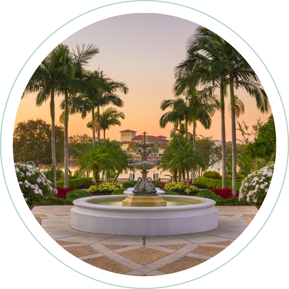 A serene outdoor scene at sunset with a fountain, palm trees, colorful flowers, and a building in the background.