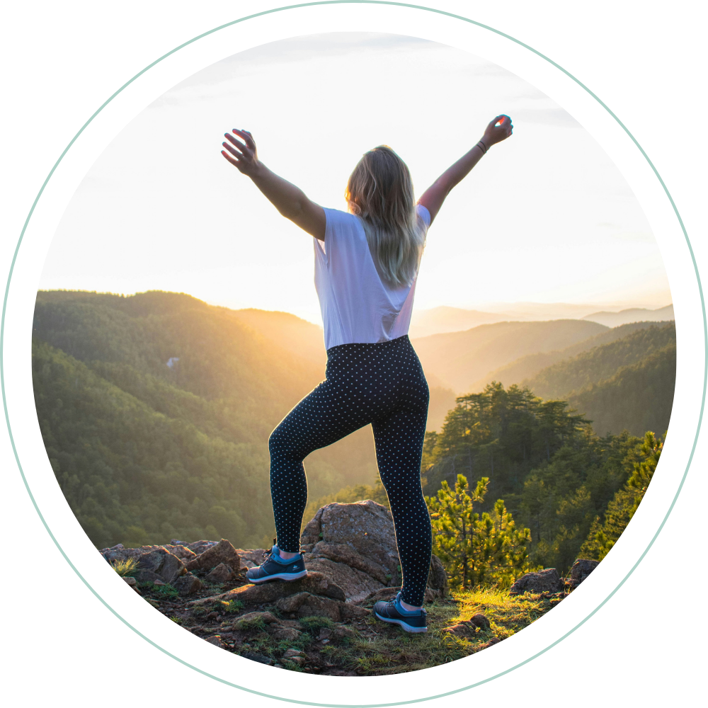 A woman with her arms raised standing on a rock at sunset overlooking a valley and mountain range.