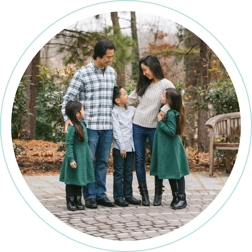 A family of six with three children, two girls and one boy, standing outdoors on a cobblestone path in a forested area during fall. The parents are smiling and looking at each other, with the mother and girls wearing green dresses and the father and boy wearing casual shirts and jeans. The scene appears warm and joyful.