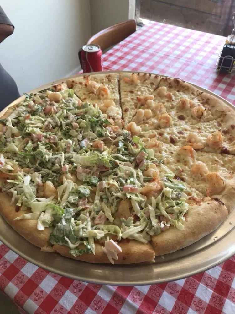 Large pizza with half topped with shrimp and cheese, and half with lettuce, bacon, and shredded chicken, on a metal tray on a red and white checkered tablecloth.