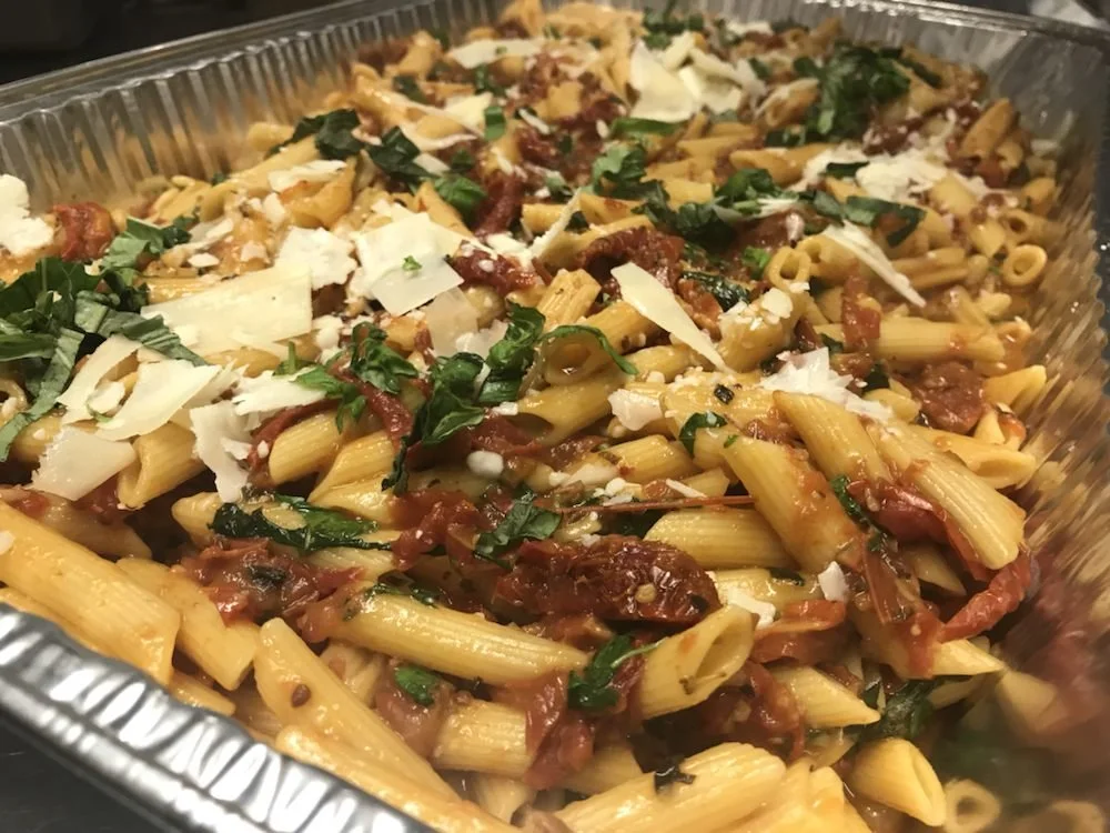 Pasta bake with rigatoni, tomato sauce, shredded cheese, chopped herbs, and sun-dried tomatoes in a foil container.