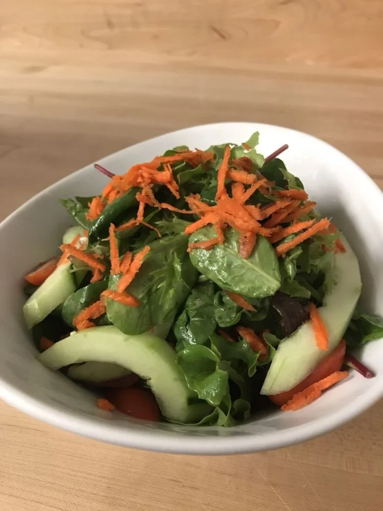 Mixed green salad with shredded carrots, sliced cucumbers, tomatoes, and leafy greens in a white bowl on a wooden surface.