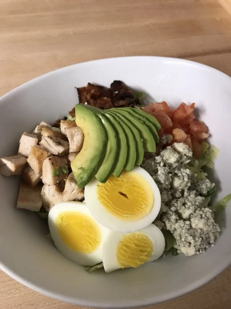Bowl of salad with sliced avocado, boiled egg, cooked chicken, crumbled blue cheese, diced tomatoes, bacon bits, and chopped lettuce on a wooden table.