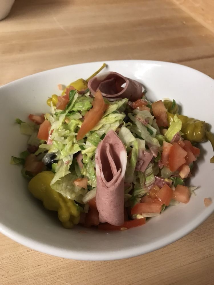 Bowl of salad with chopped lettuce, diced tomatoes, yellow peppers, shredded ham, sliced black olives, and rolled slices of deli meat, on a wooden table.