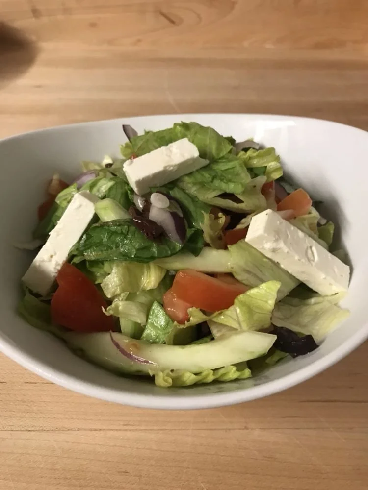 A bowl of Greek salad with lettuce, tomatoes, red onions, black olives, and feta cheese on a wooden surface.