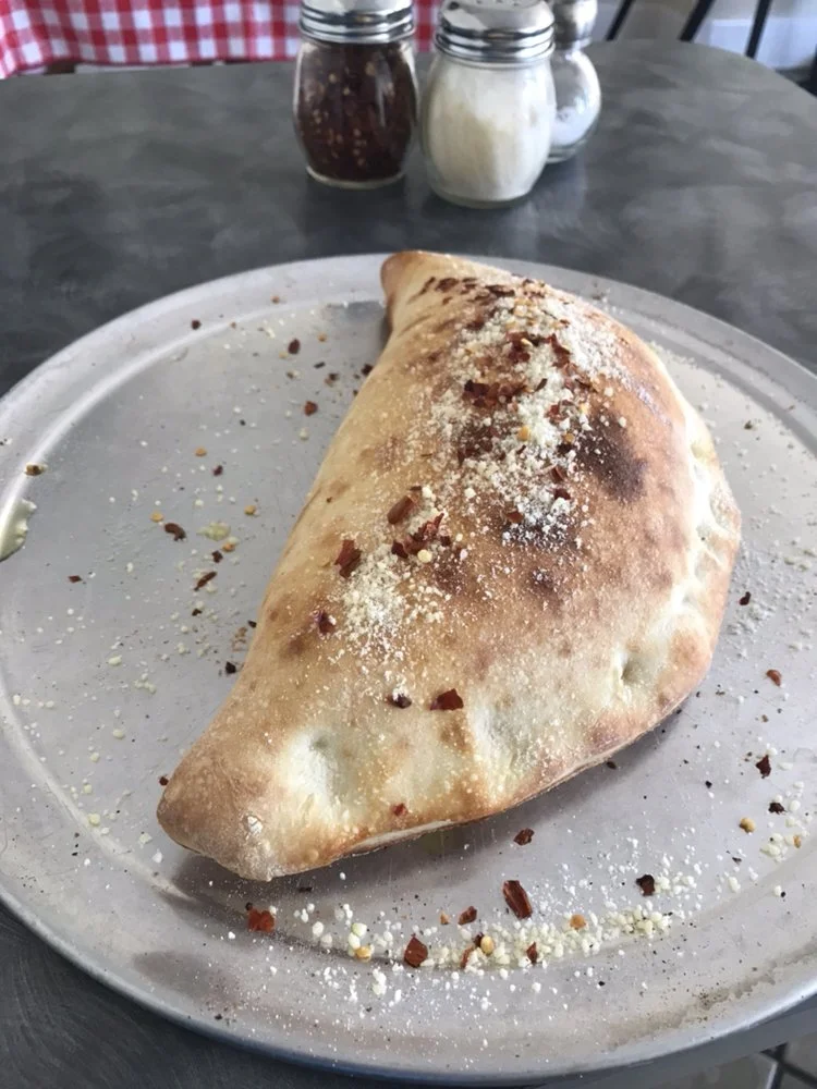 A calzone-style folded pizza with cheese and toppings, sprinkled with crushed red pepper and grated cheese, on a round plate.