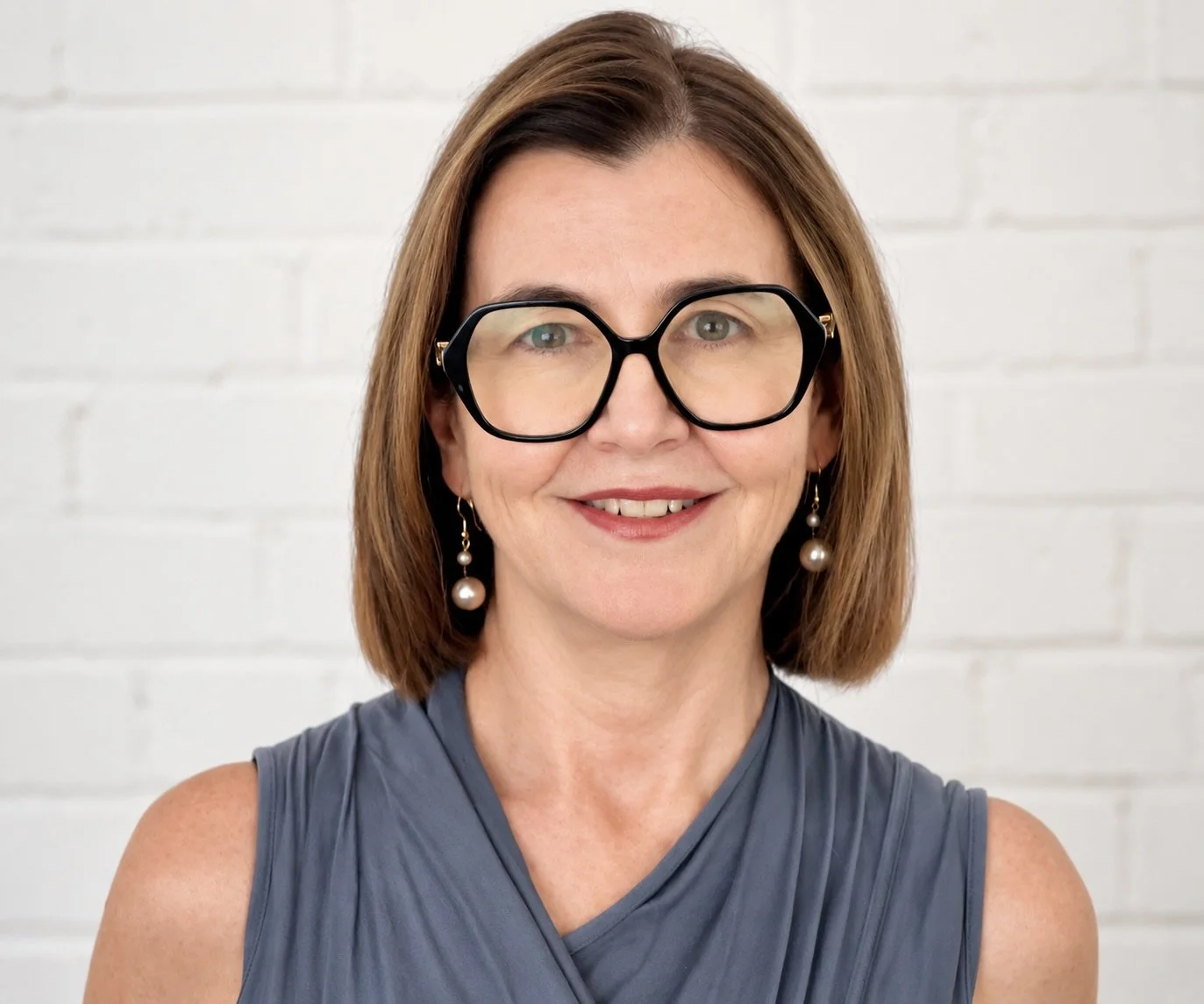 A woman with shoulder-length brown hair, wearing large black glasses, pearl earrings, and a sleeveless blue top, standing against a white brick wall.
