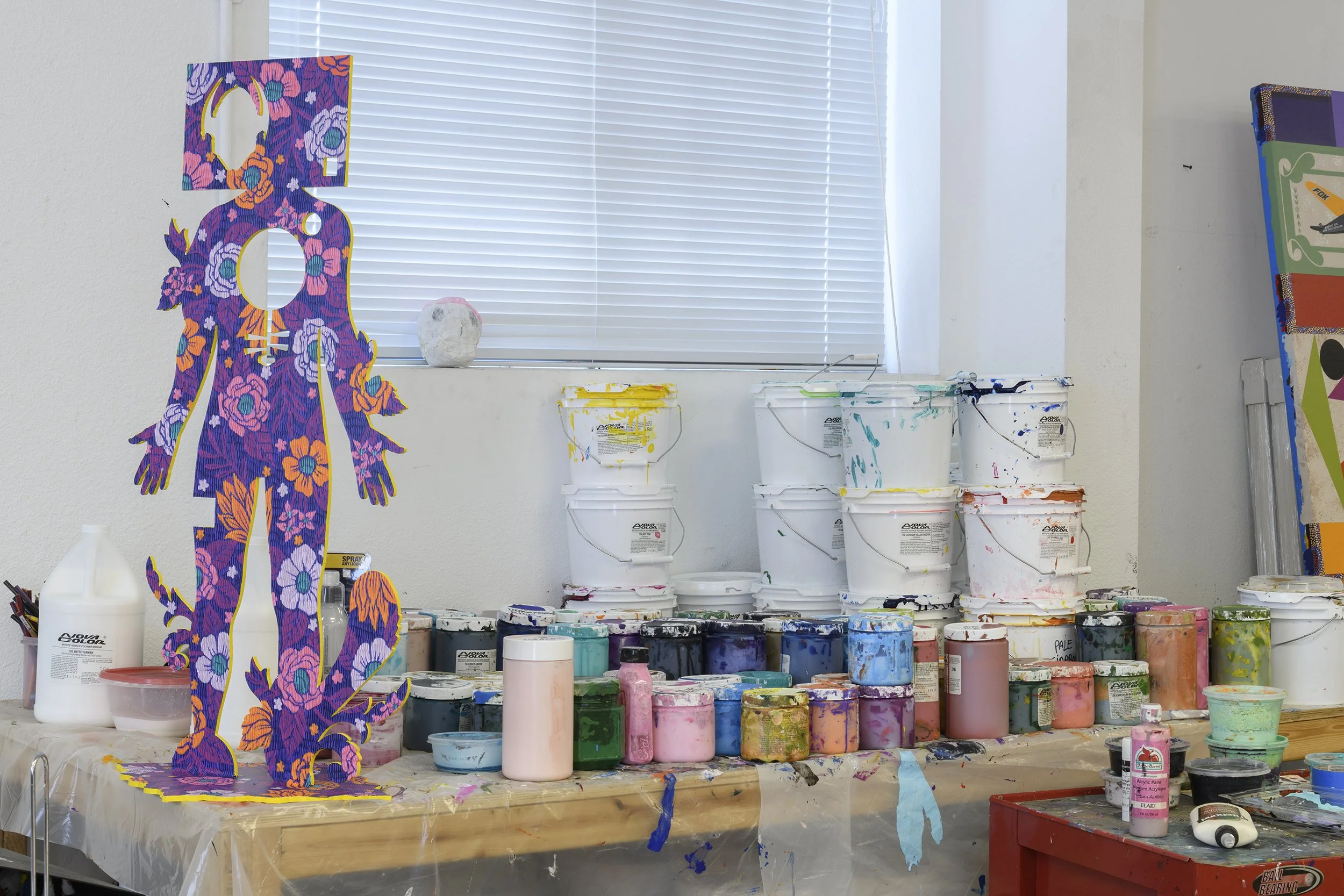An art studio workspace with a colorful, floral manikin figure cutout on the left, and stacks of white, paint-splattered buckets behind a table covered with bottles of paint and art supplies. A window with closed blinds is in the background.