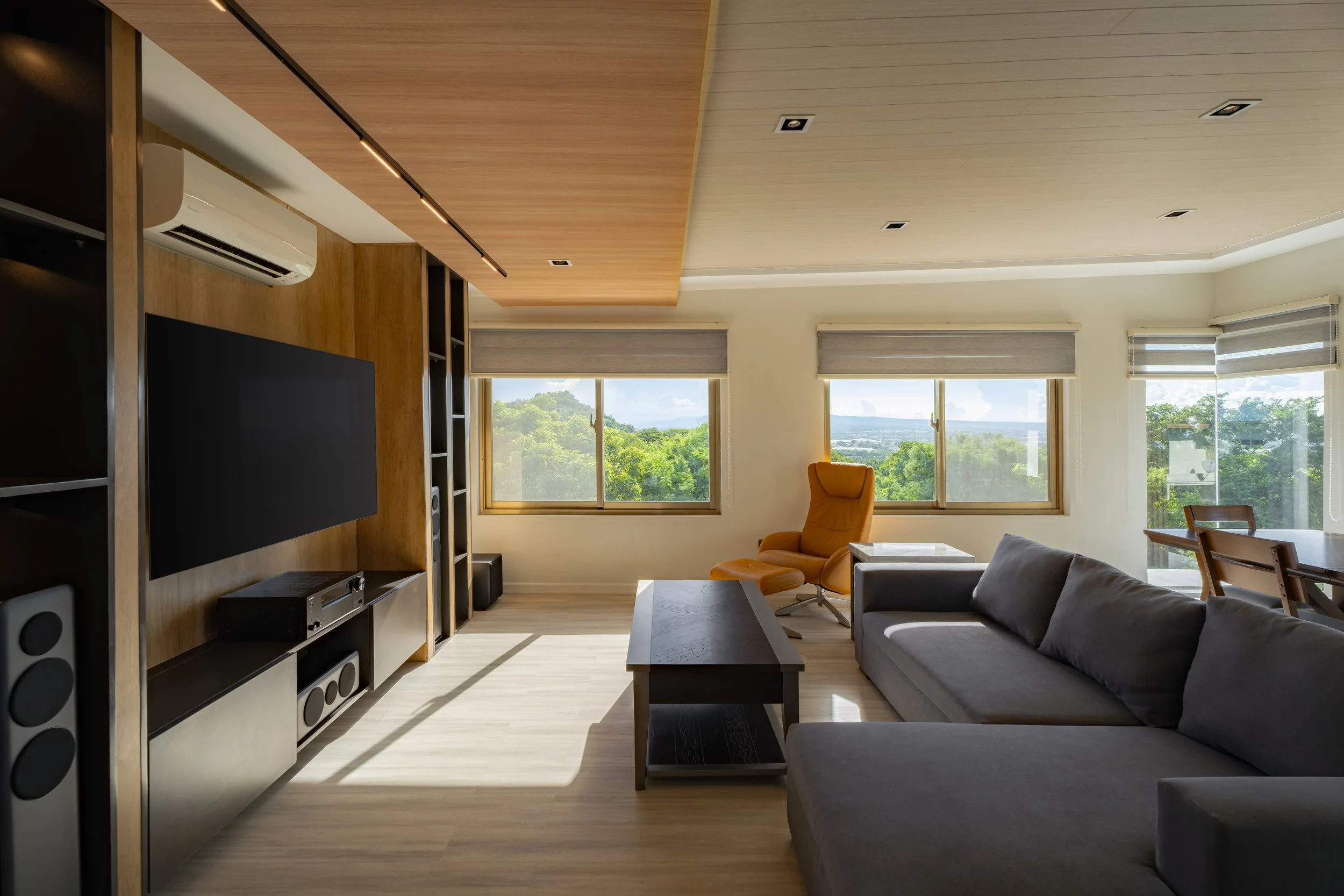 Modern living room with large windows showing greenery outside, gray sofa, yellow armchair, black coffee table, wooden ceiling, and entertainment center with TV and speakers.