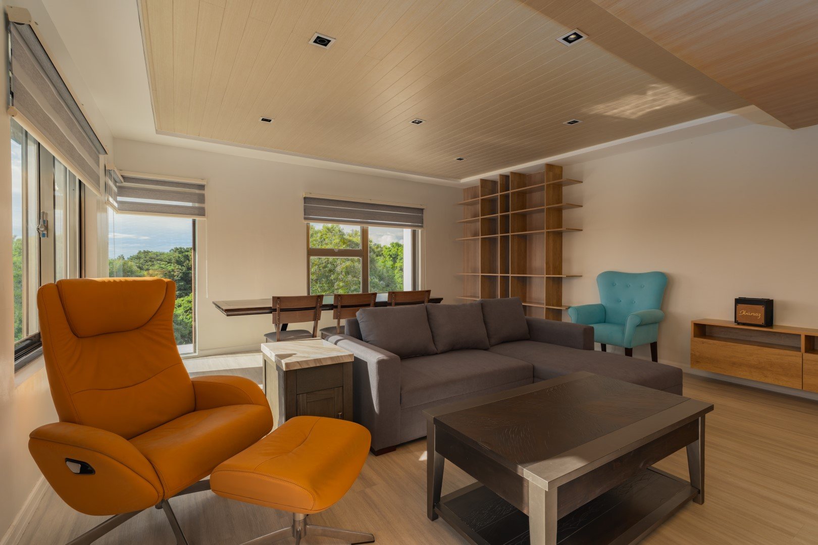 Living room with orange swivel chair, gray sofa, wooden shelving unit, blue armchair, wooden side table, and large windows showing greenery outdoors.
