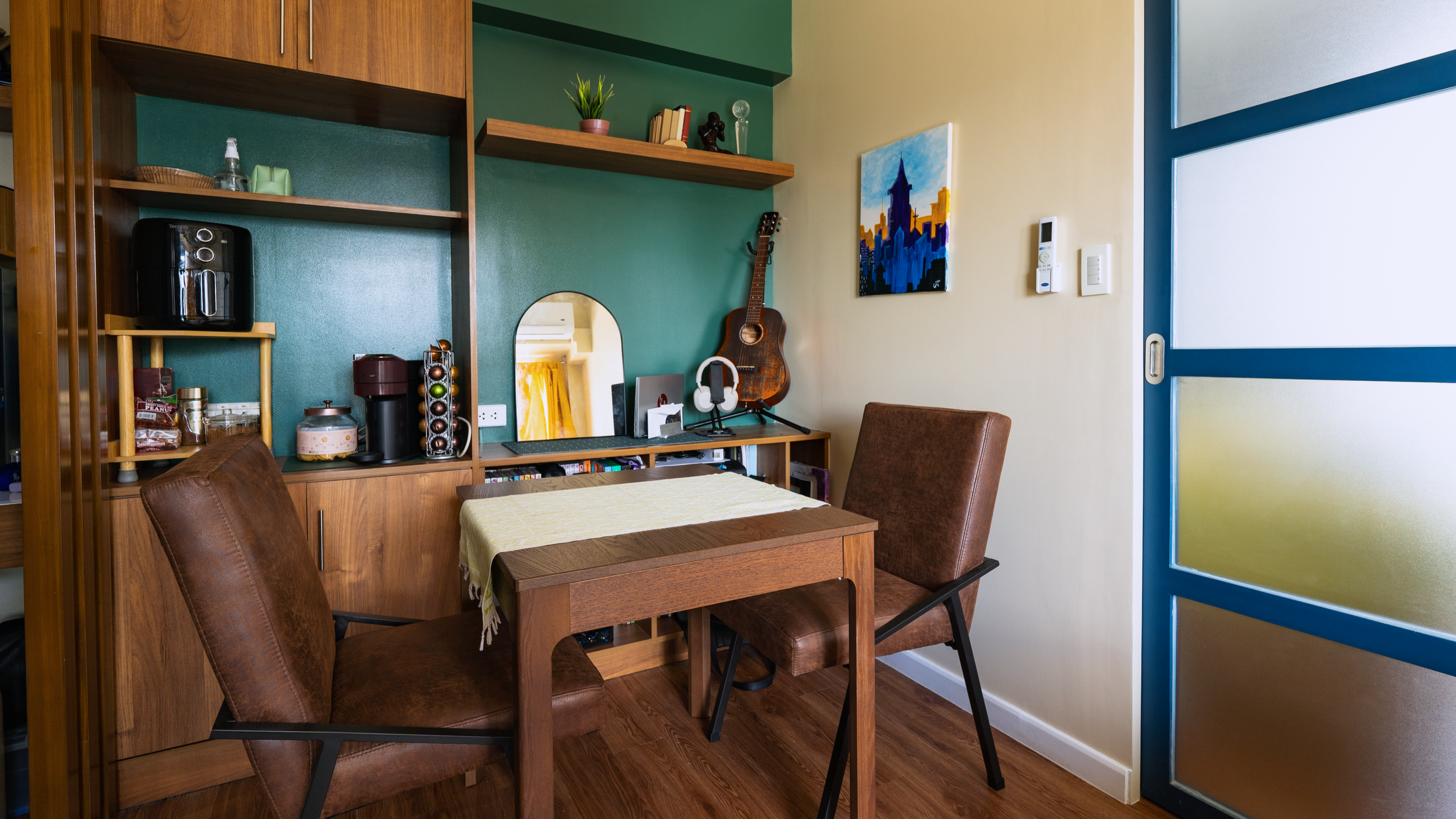 A cozy dining area with a wooden table and brown leather chairs. Behind the table is a wooden cabinet with open shelves holding various items including a coffee machine, jars, books, and decorative objects. A framed mirror and an acoustic guitar are also on the cabinet. The wall is painted two shades of green, and a colorful cityscape painting is hung on the wall. A sliding door with frosted glass and blue frame is on the right side of the image.