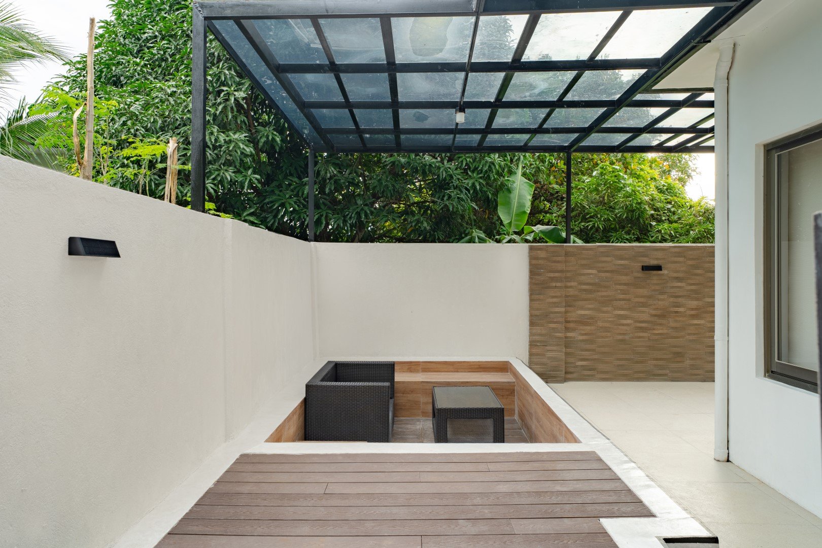 A modern outdoor patio area with a glass roof, white and beige walls, greenery in the background, and black outdoor furniture including a sofa and a small table. There are wall-mounted lights on the walls.