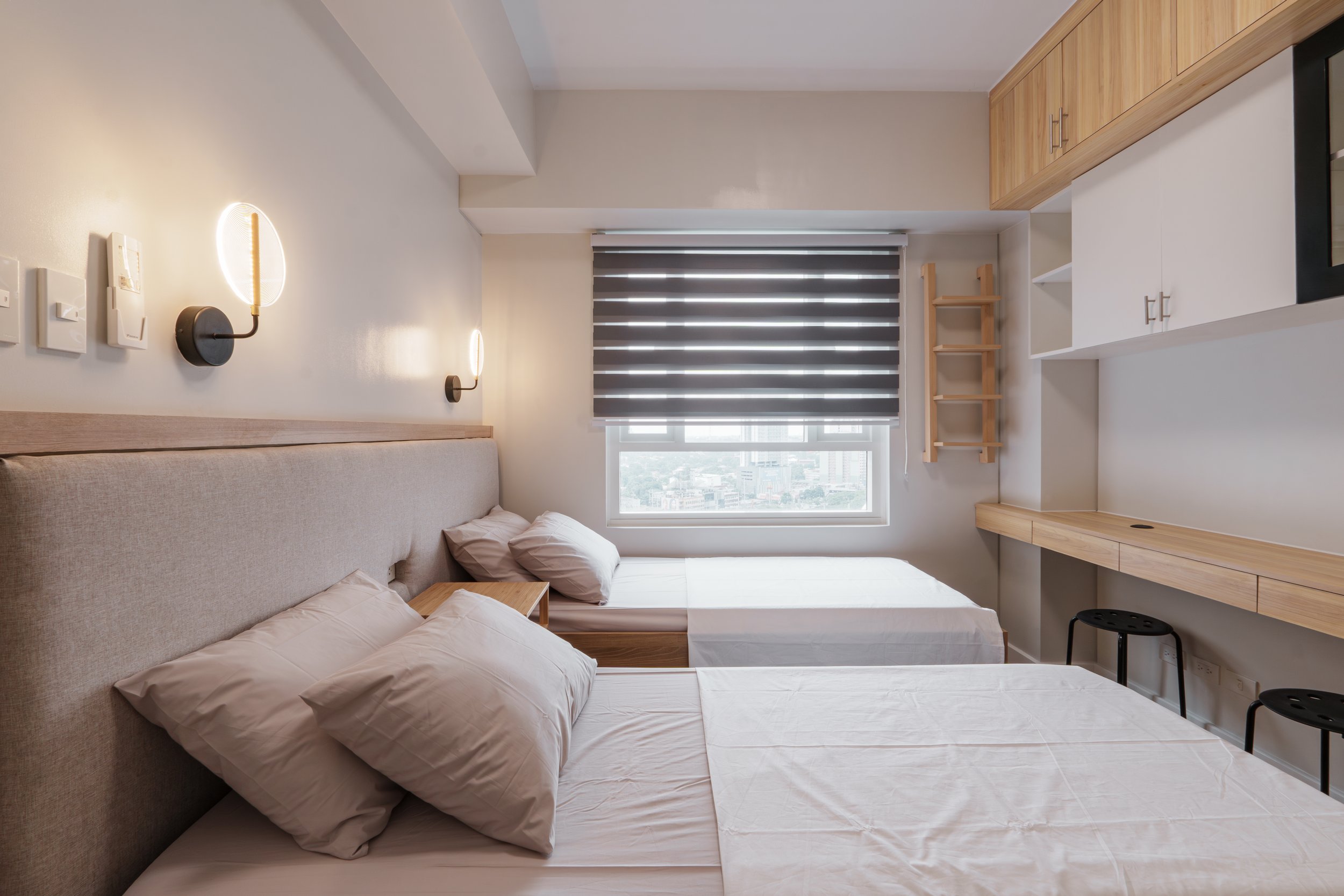 A bedroom with two beds with beige headboards and white bedding, a window with black and white striped curtains, wooden furniture, and two wall-mounted lamps.