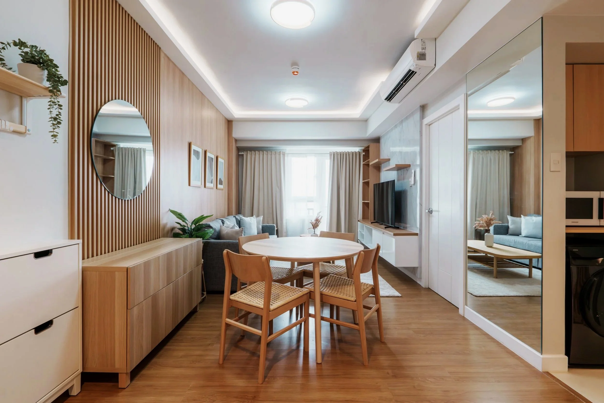 Modern living room with wooden accents, a round dining table with four chairs, a sofa, wall-mounted TV, and a large mirror reflecting part of the room, with natural light coming through windows covered with light curtains.