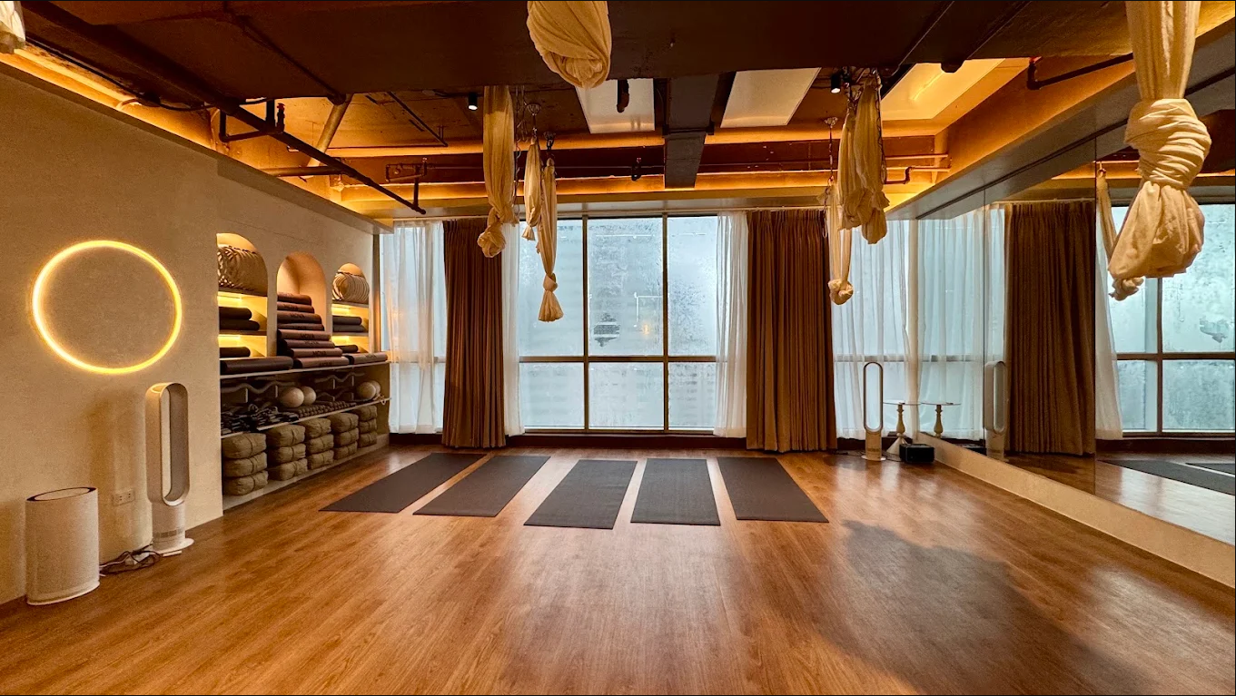 Empty yoga studio with black mats on wooden floor, large windows with curtains, hanging fabric swings, and shelves with rolled towels and fitness balls.
