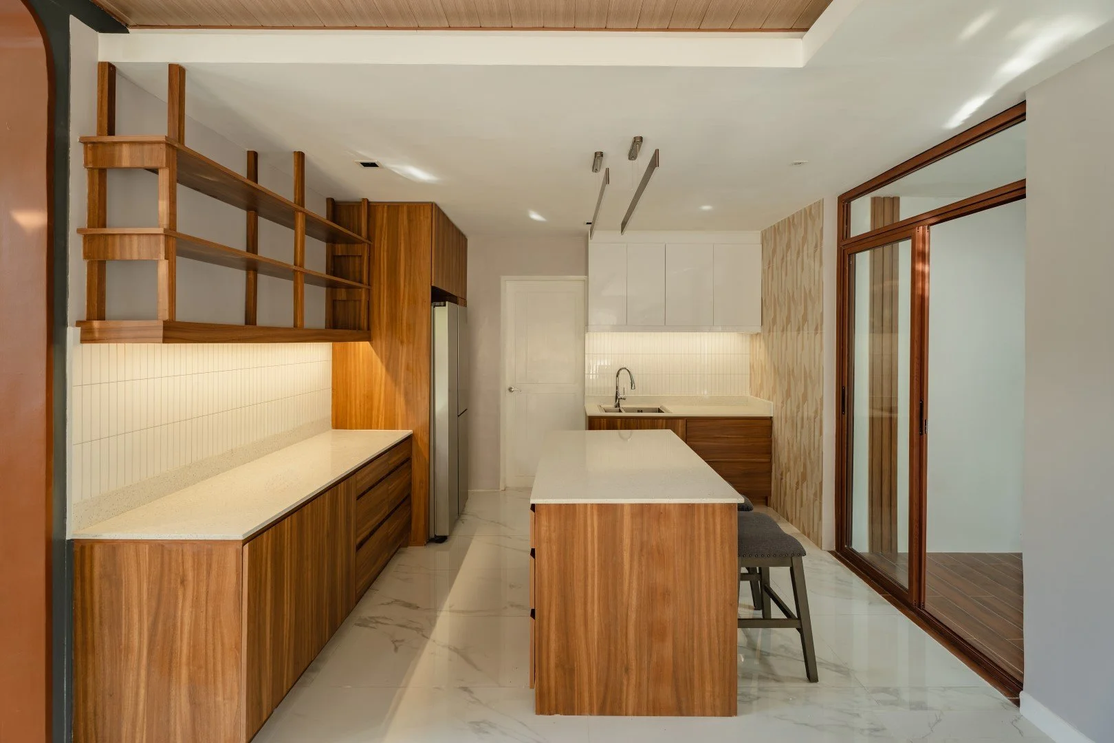 Modern kitchen with wood and white cabinetry, marble countertops, a stainless steel refrigerator, and a sliding glass door leading to an outdoor area.