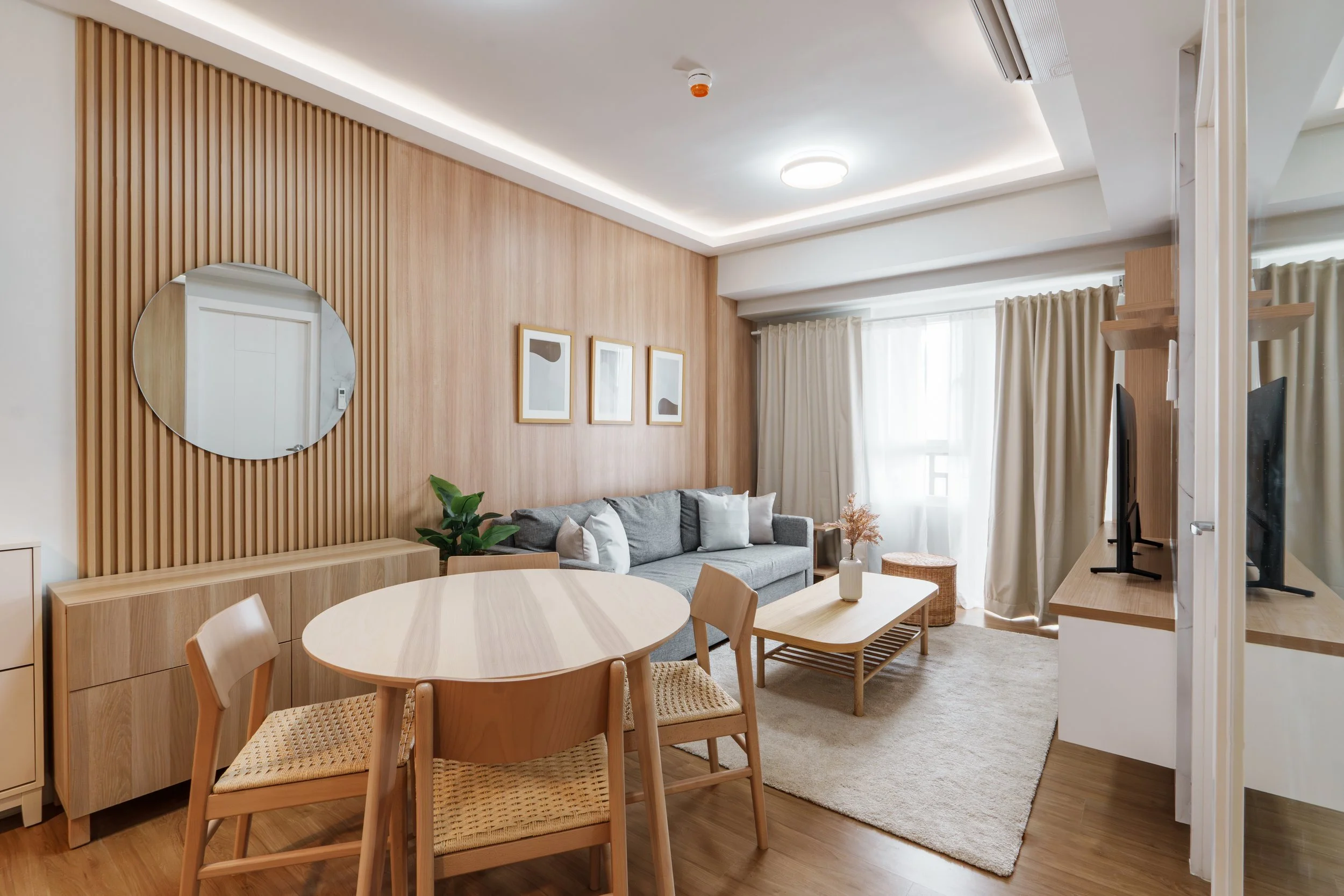 Modern living room with a dining table, wooden chairs, a gray sofa, a TV, and decor with natural light from large windows and beige curtains.