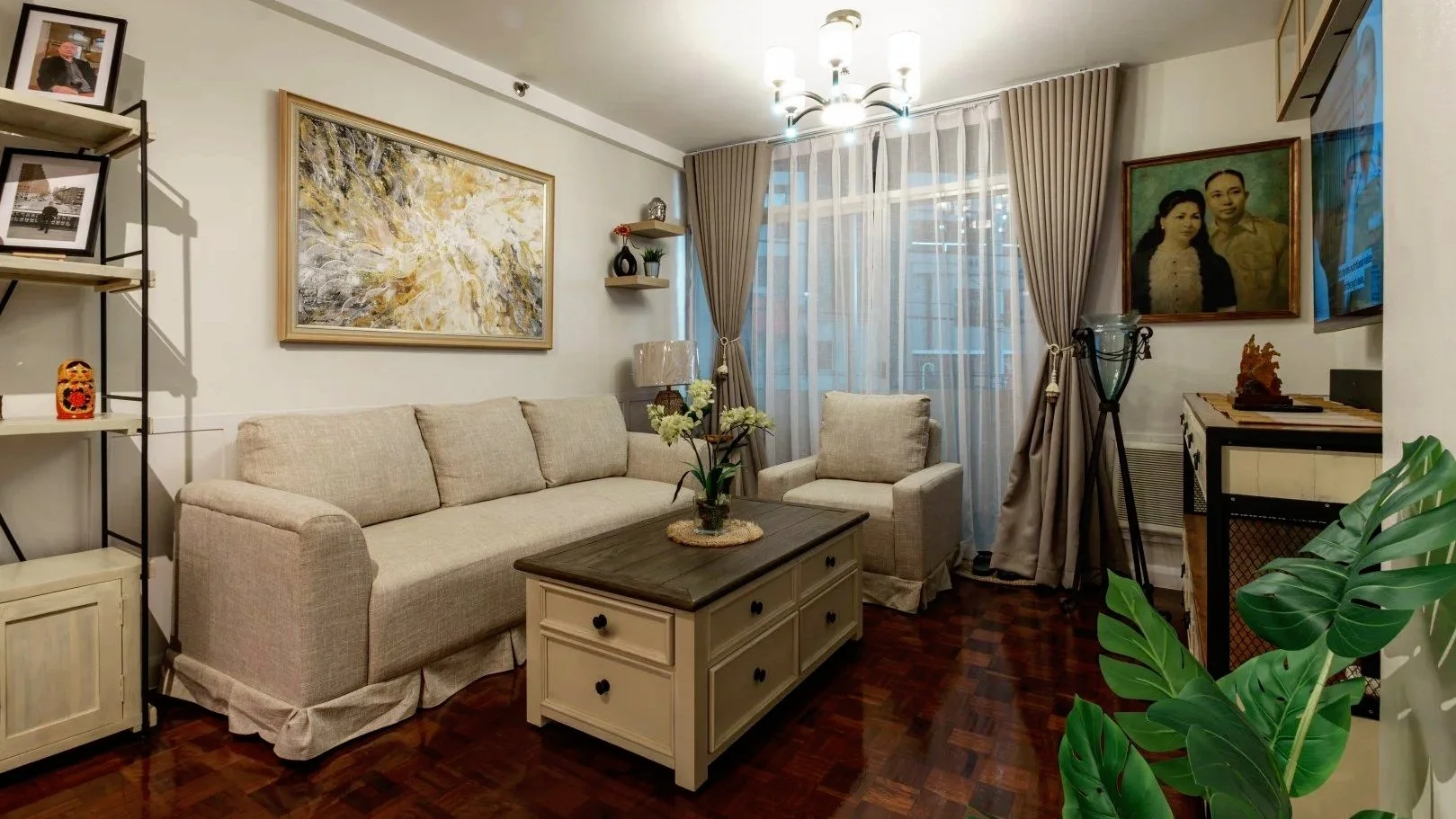 A cozy living room with a beige couch and armchair, a dark wood coffee table with floral decor, a bookshelf with framed photos and a decorative jar, a large abstract wall art, window with beige curtains, a floor lamp, framed family portrait, a flat-screen TV, a potted plant, and decorative shelves and sculptures.