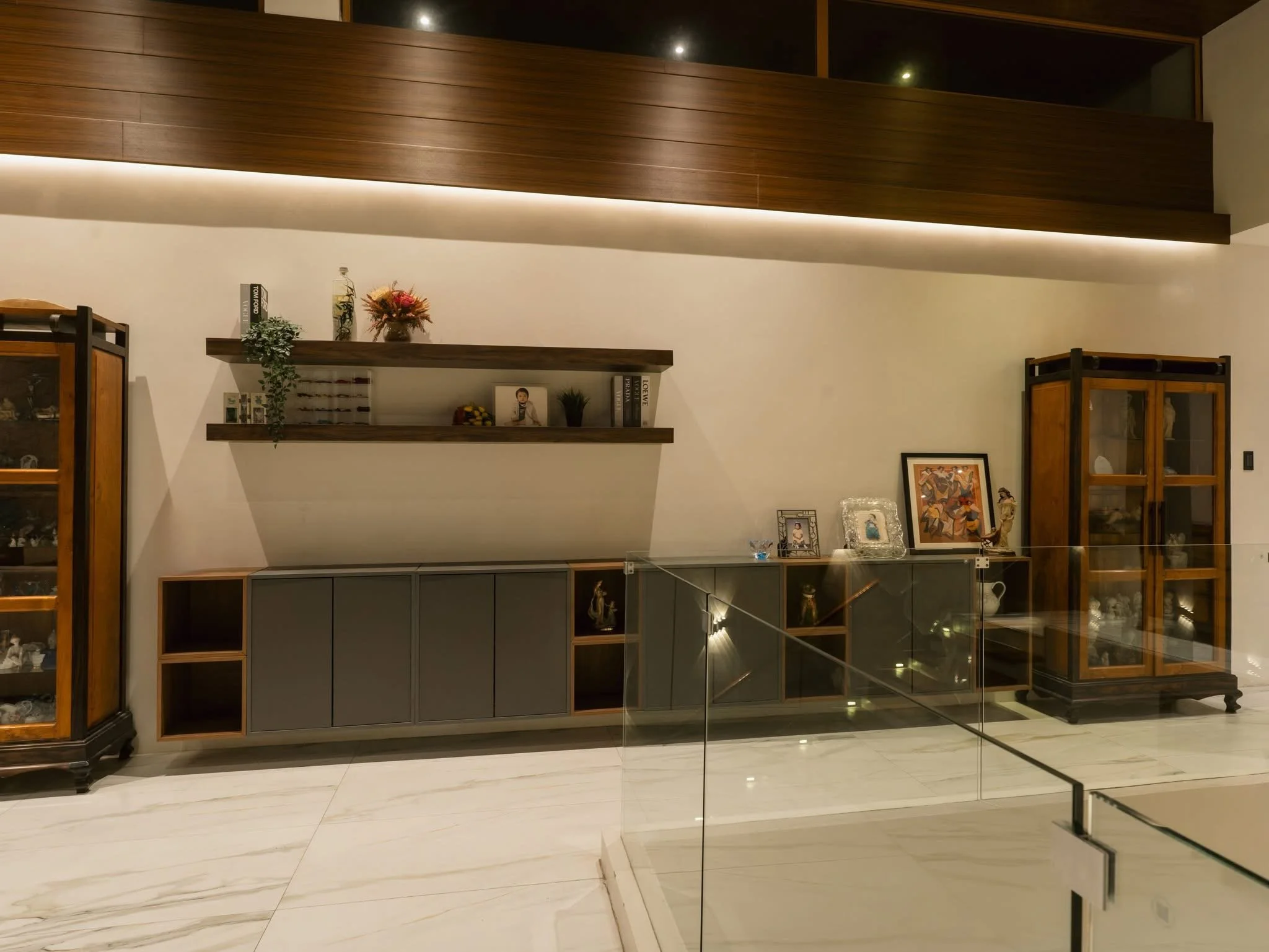 Interior view of a modern living space featuring wooden and glass display cabinets, framed artwork, decorative items, and floating wall shelves with books and flowers, illuminated by warm lighting.