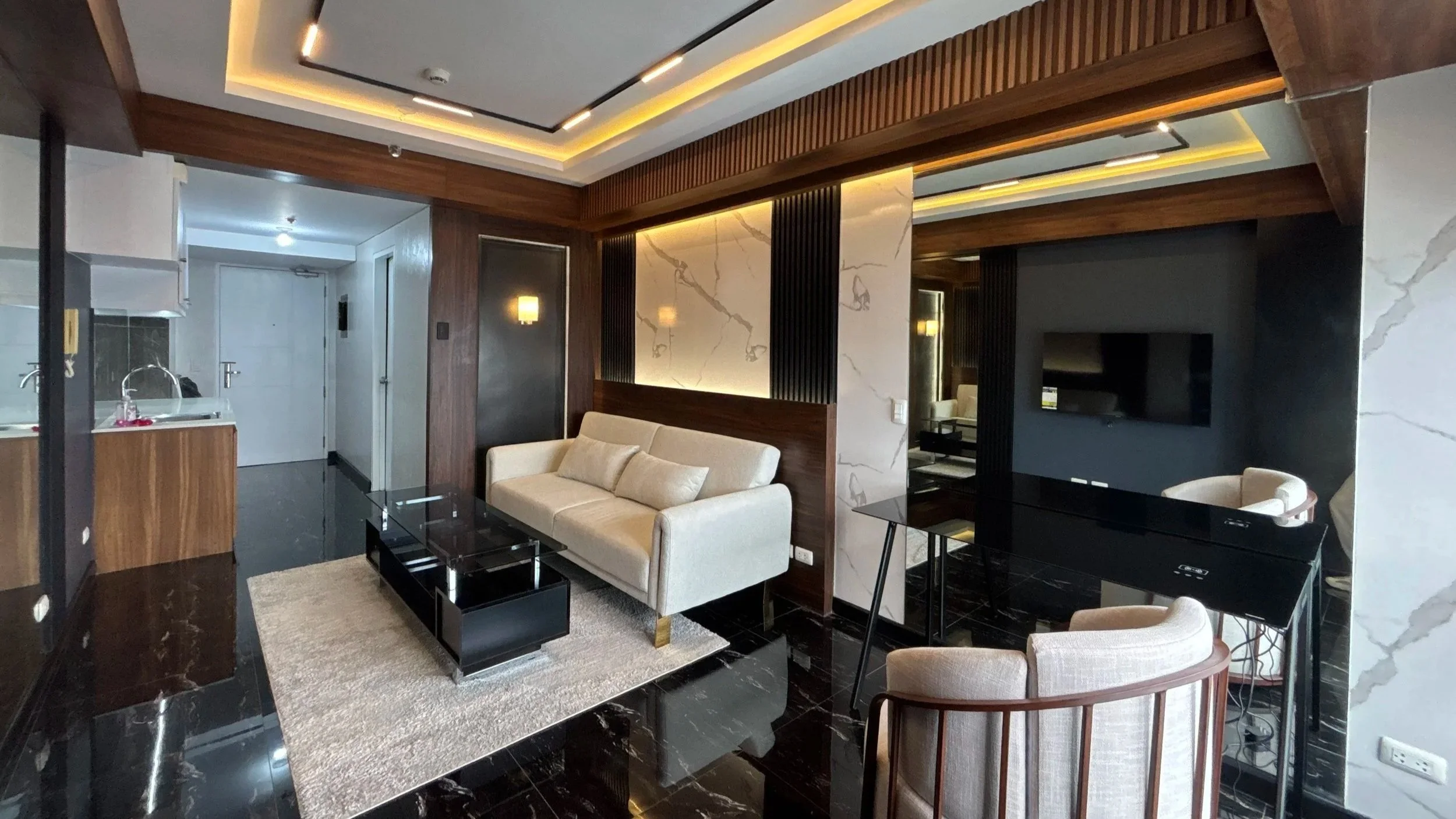 Modern living room with beige sofa, glass coffee table, black TV stand, wall-mounted TV, white marble accent wall, wood panel accents, and a black and white marble floor.