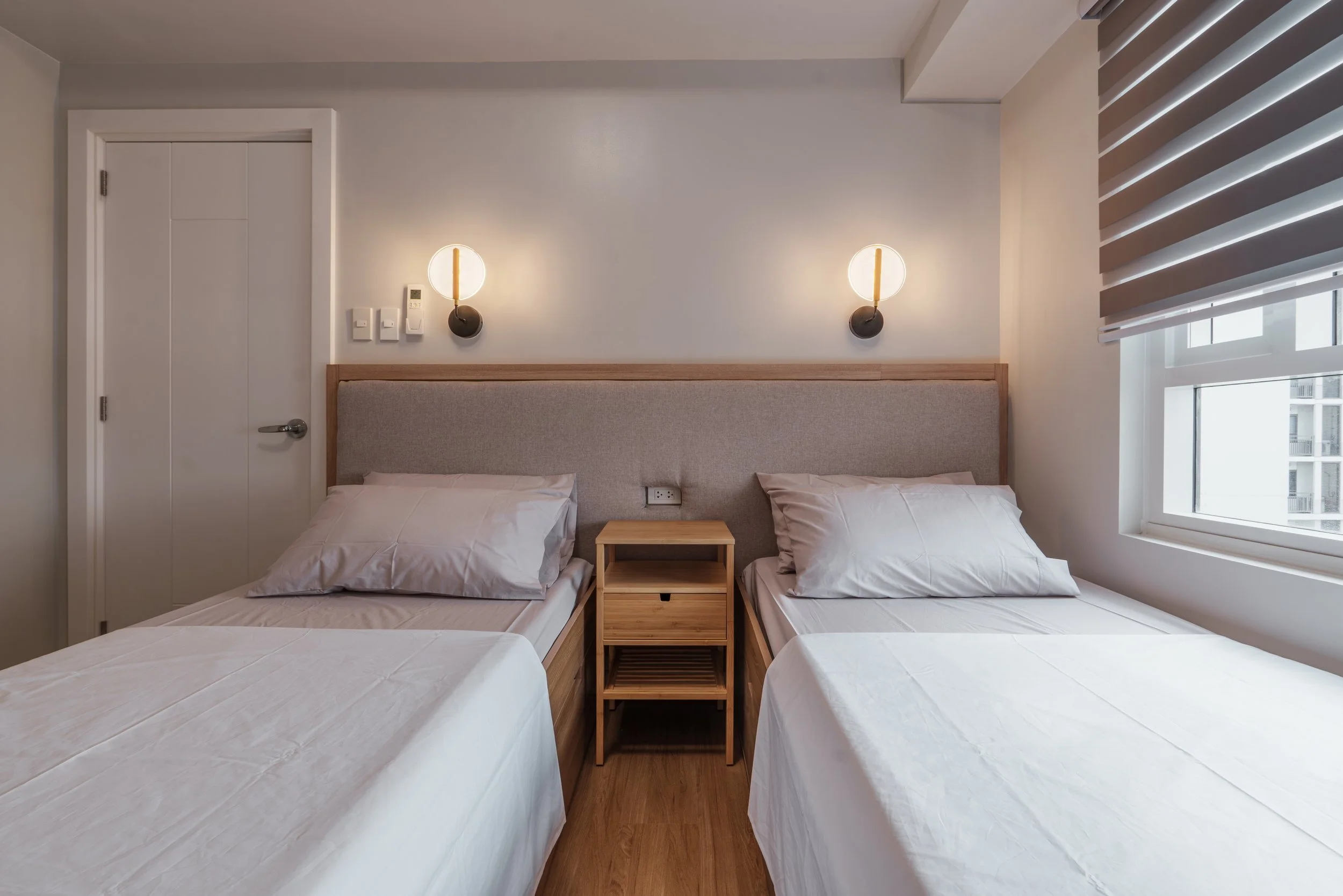 A modern hotel room with two single beds separated by a wooden nightstand, white bedding, and a padded headboard. Two wall-mounted lamps and a window with horizontal blinds are also visible.