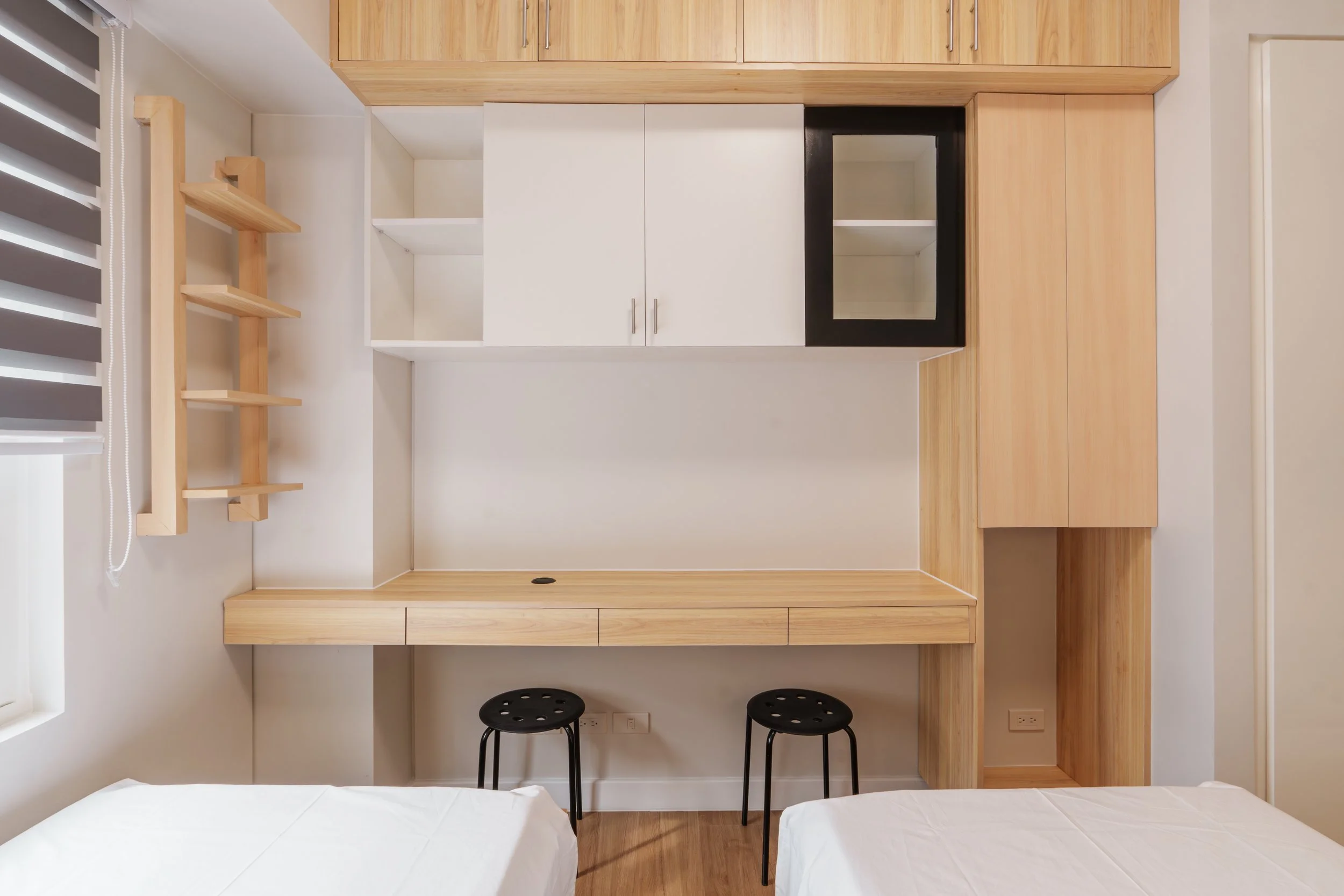 Empty bedroom with a modern wooden and white built-in wall unit, two black stools, and two beds with white sheets.