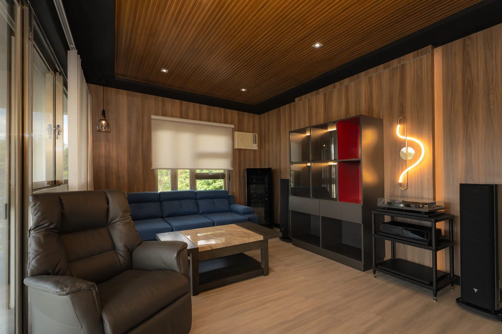 Living room with wood-paneled walls and ceiling, featuring a brown leather recliner, a blue sofa, a wooden coffee table, a large window with a white roller blind, and a modern entertainment center with decorative lighting, speakers, and electronic equipment.