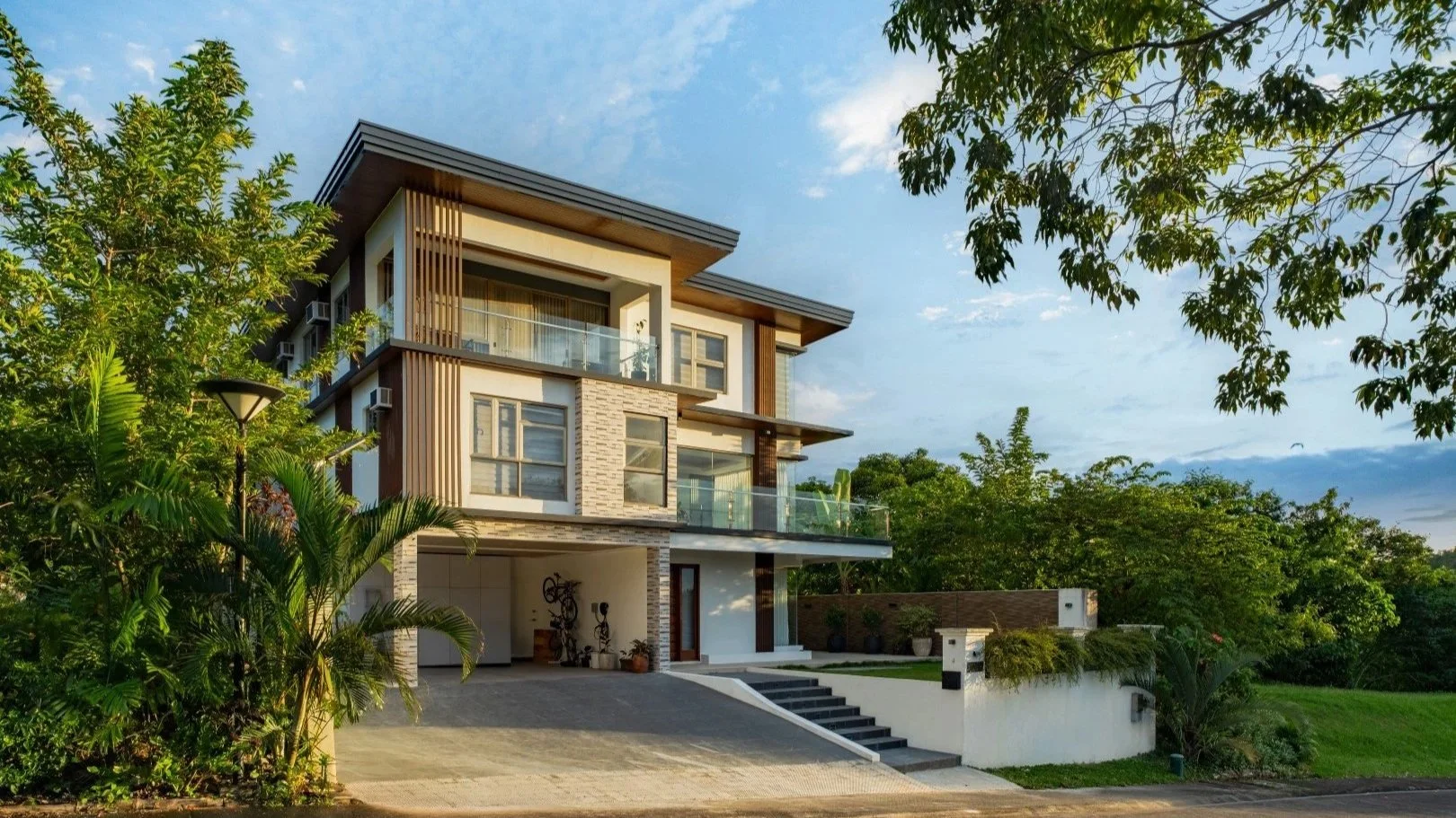 Modern multi-story house with large windows, balconies with glass railings, surrounded by greenery and trees, under a blue sky with scattered clouds.