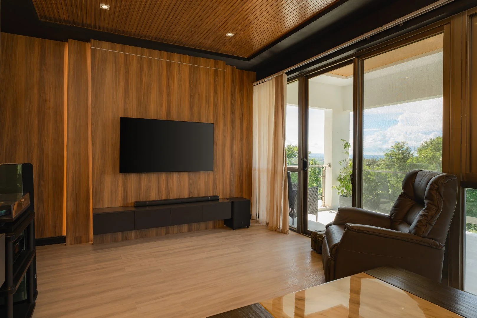 Living room with wood-paneled wall, mounted TV, sliding glass door leading to balcony, and a brown leather recliner facing outside.