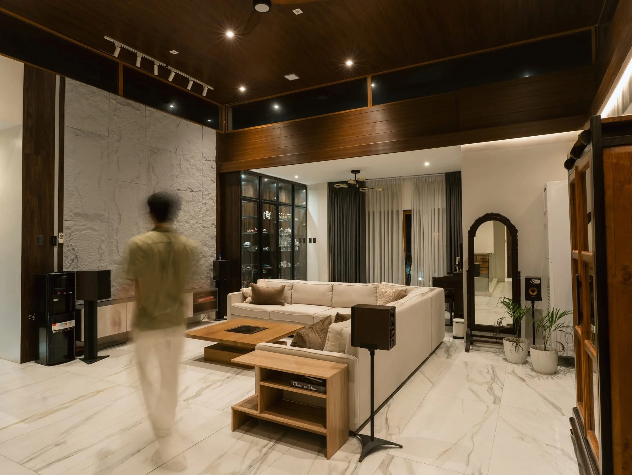 A modern living room with a white sectional sofa, a wooden coffee table, and a black display cabinet with glass doors. The room has white marble floors, textured white stone walls, dark wooden ceiling, recessed lighting, and large windows with curtains. A blurred person is walking past.