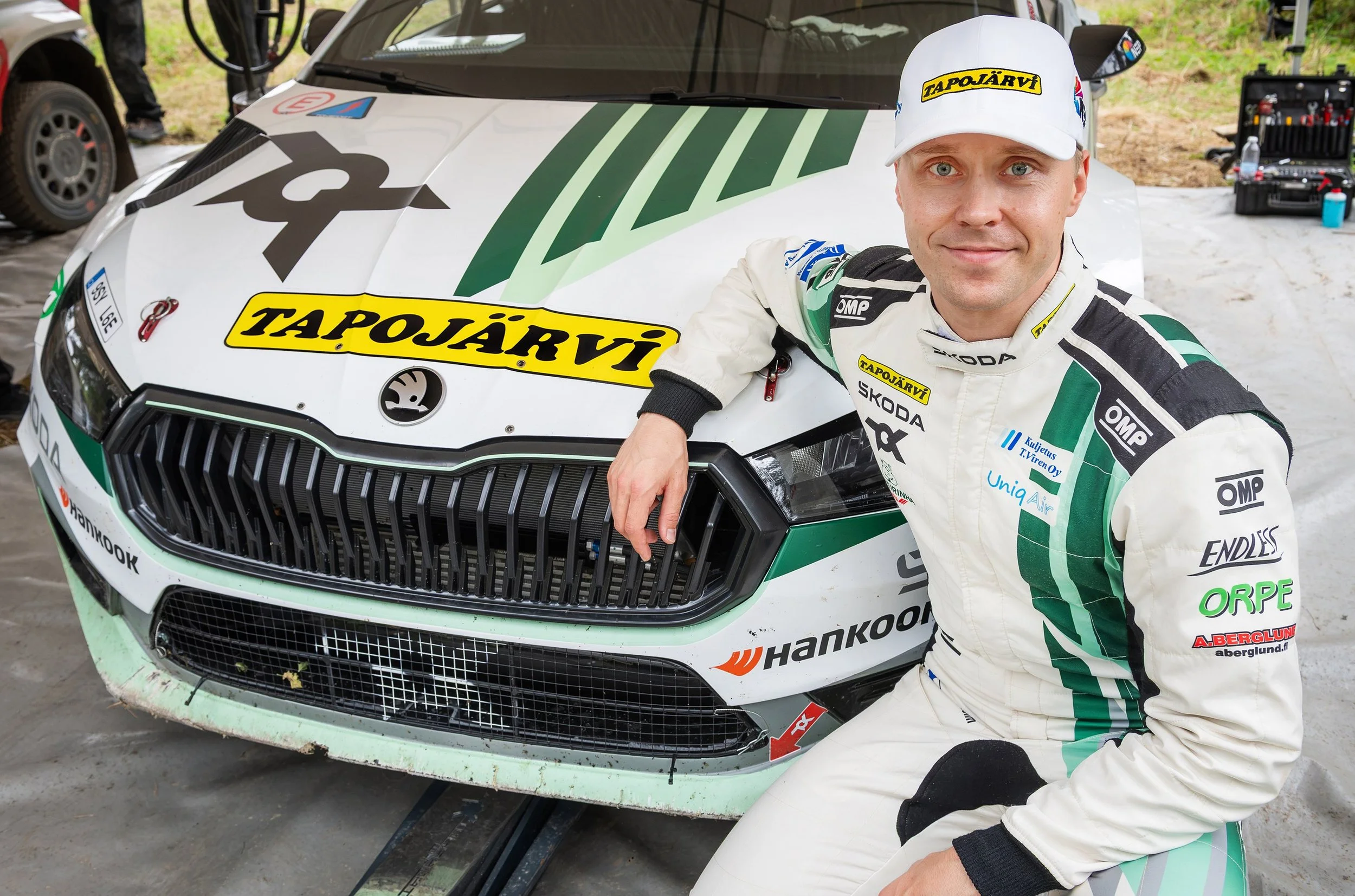 A race car driver wearing a white racing suit and cap standing next to a white rally car with various sponsor logos. The driver is smiling and has his arm resting on the car.