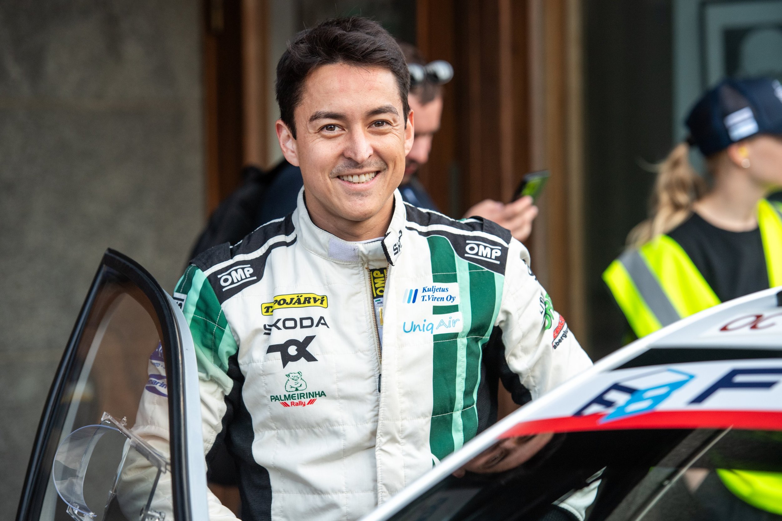 A smiling co-driver in a white and green racing suit standing outside, with other people and a racing vehicle around him.