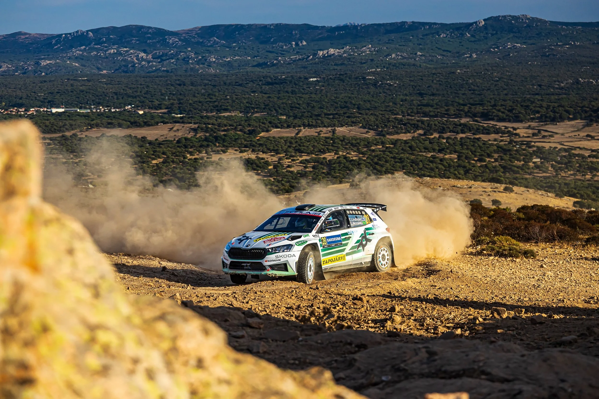 A rally car racing on a dirt road in a mountainous landscape, kicking up dust.