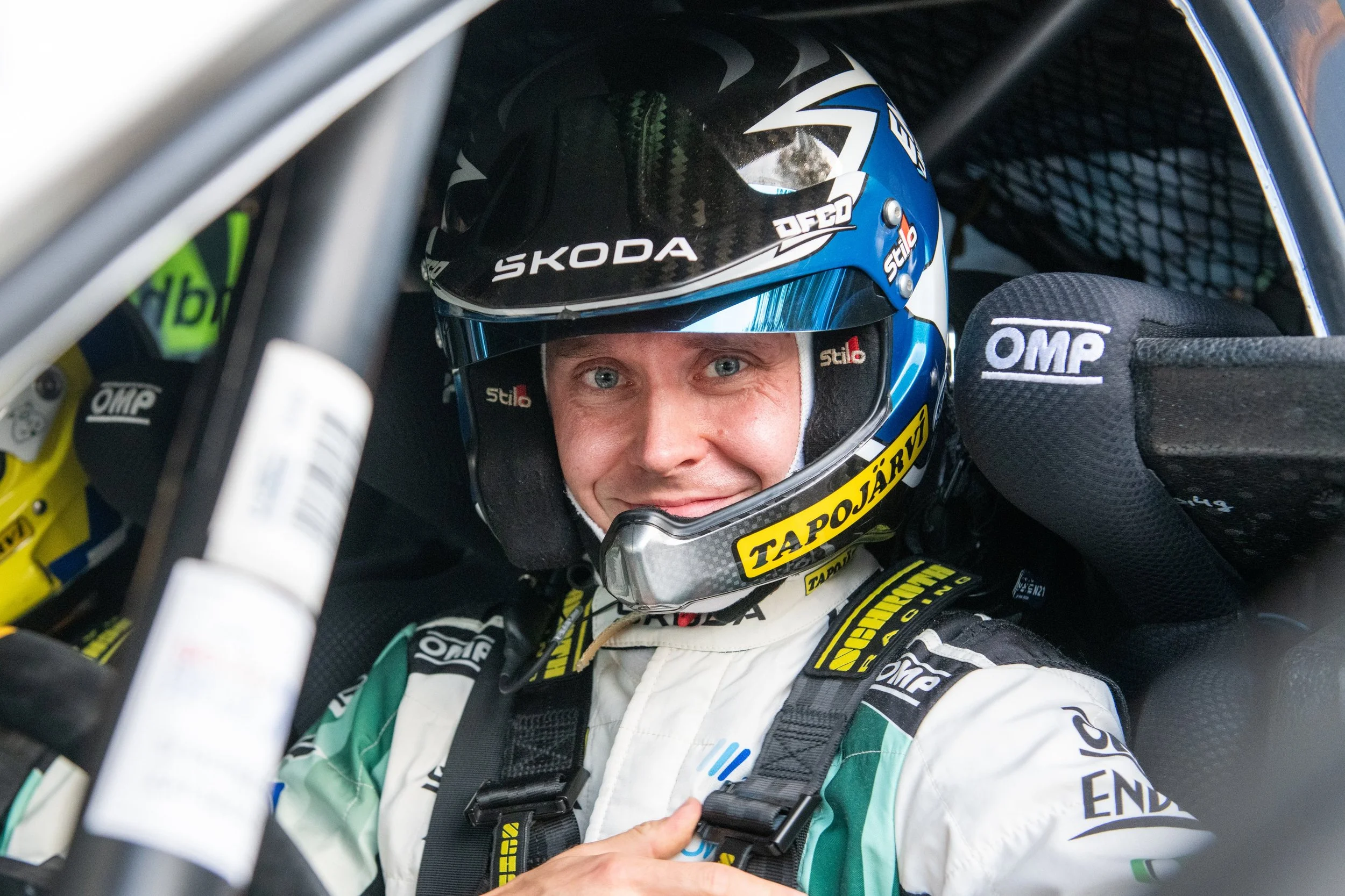 Close-up of race car driver sitting in car, wearing blue and black helmet with sponsor logos, white racing suit with sponsor patches.