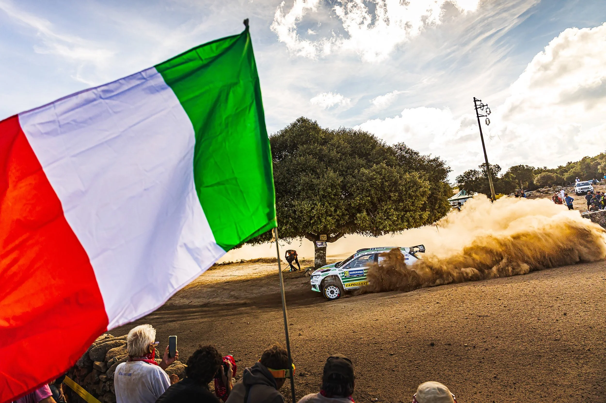 An Italian flag waves in the foreground beside a rally car racing on a dirt track. Spectators watch from the sidelines as the car kicks up dust during a daytime motorsport event, with trees and a cloudy sky in the background.