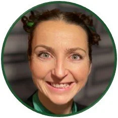 Nina  in close-up portrait, likely for personal or professional identification. She wears green neckwear and hair accessories, with dark clothing against a softly blurred gray background.