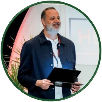 Arad  holding a tablet while presenting onstage. Red accent lighting, tall plant, projection screen, and event setting frame speaker.